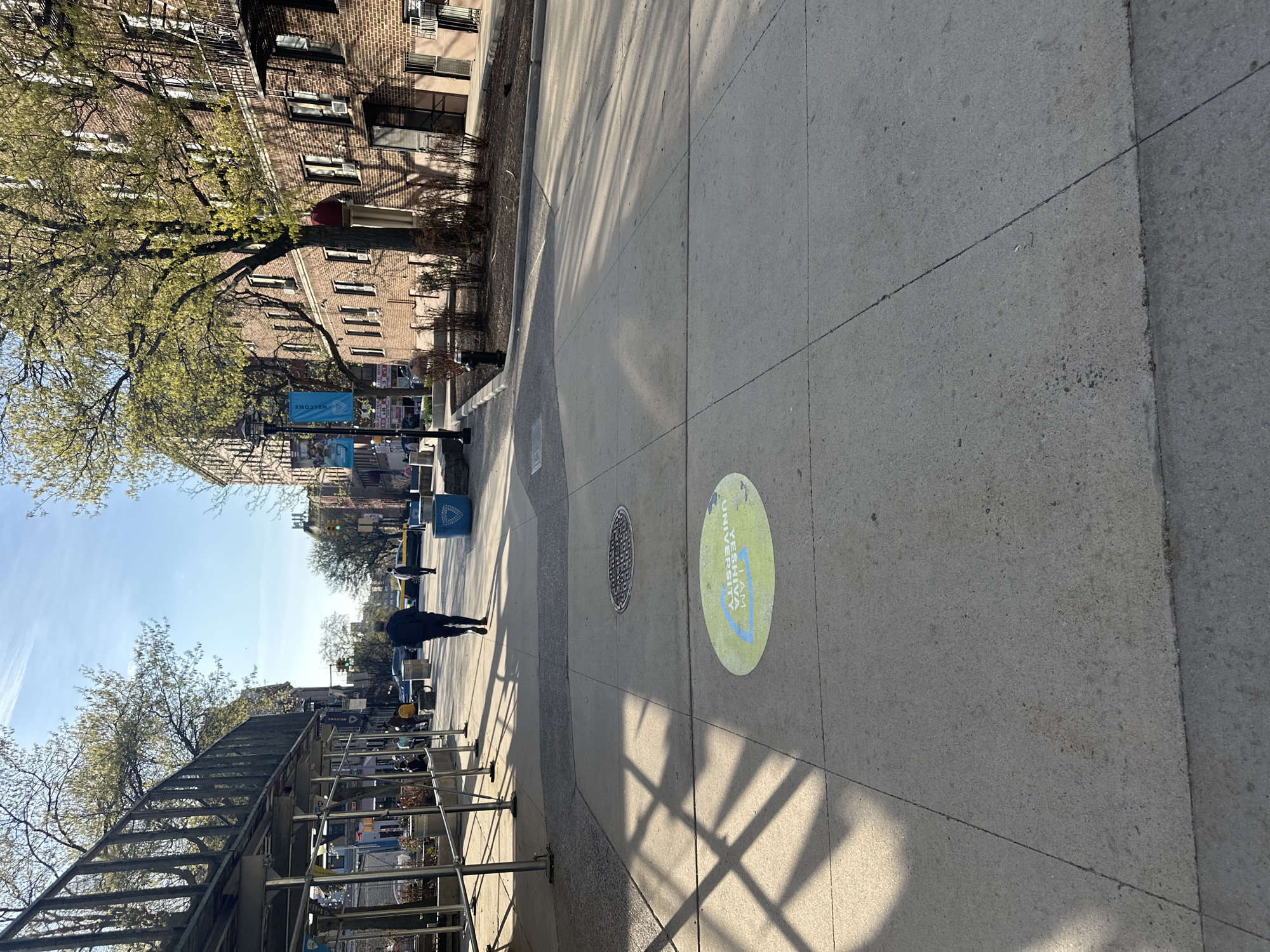 The image depicts a sidewalk in New York City, United States of America. The sidewalk is made of concrete slabs. Shadows of trees and scaffolding cast dark lines across the surface. A green circular sticker on the ground reads "I AM YIM UNIVERSITY." A manhole cover is visible nearby. Scaffolding is set up along the left side of the sidewalk. Buildings and trees line the left side of the street. A person in a dark coat walks in the distance, heading away from the viewer. Banners hang from lampposts along the street.