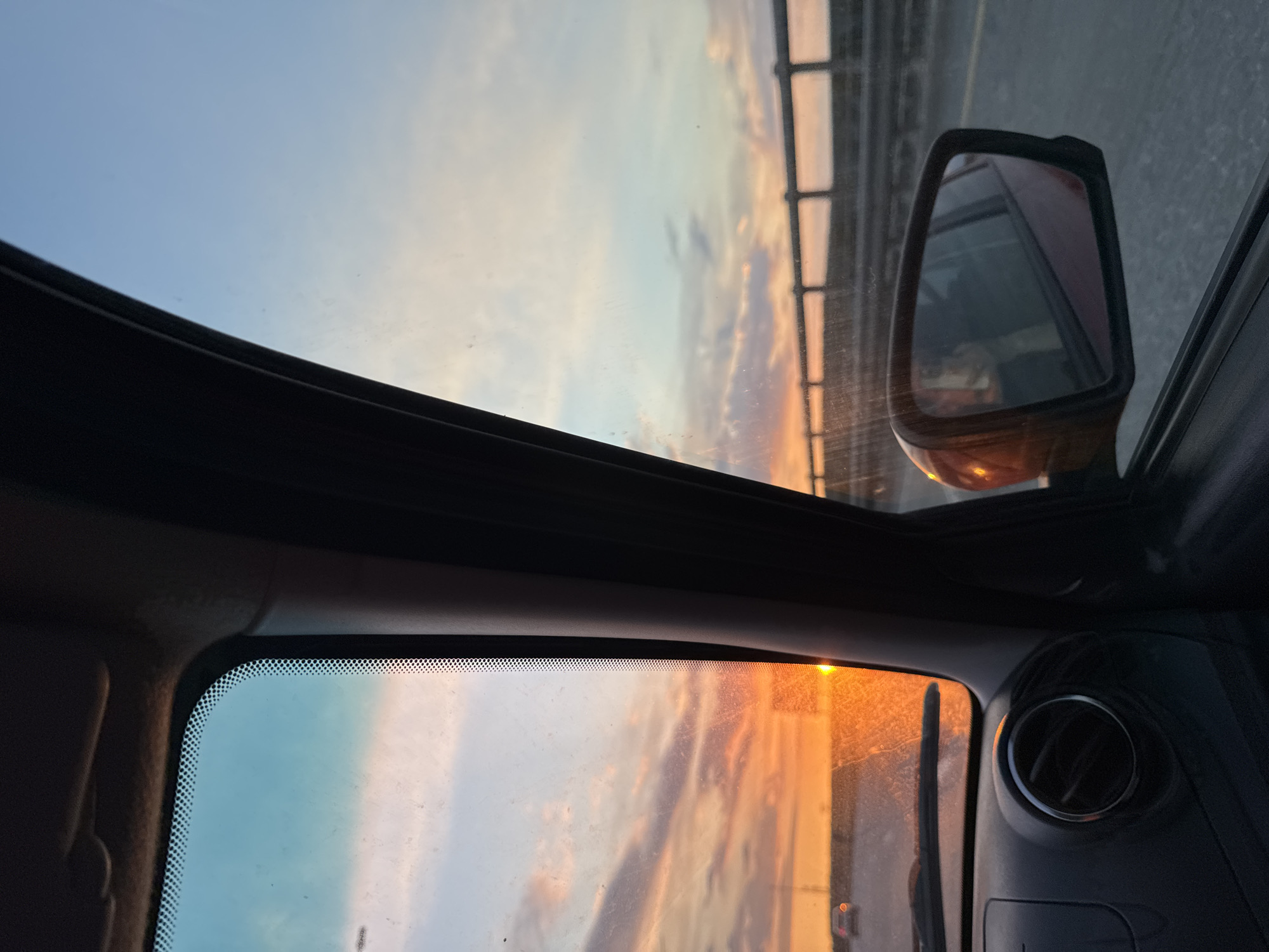 The image presents a serene view from inside a vehicle, looking outwards through the front windshield and partially through a sunroof, capturing the transition of light during either dawn or dusk. The sky dominates the exterior view, displaying a gradient of colors from a light, clear blue in the upper left to warm orange, yellow, and pink hues on the right, indicating the low position of the sun. Scattered clouds are visible against this colorful backdrop.
A strong, bright orange glow on the right side suggests the presence of the sun near the horizon, casting a warm radiance over the scene. Outside the vehicle, a distinct metallic structure with visible vertical and horizontal railings is seen through the side window and partially reflected in the exterior side mirror, suggesting a bridge or an overpass. The side mirror itself is dark-framed, showing a blurred reflection.
Inside the vehicle, parts of the dark dashboard are visible on the right, including a circular air vent with a chrome trim. The windshield appears to have some dust or smudges. A frit band, a pattern of small black dots, is noticeable along the top edge of the windshield. The overall atmosphere conveys a peaceful moment of travel in Perm, Russia, against a beautiful sunrise or sunset. No discernible text is visible in the image.