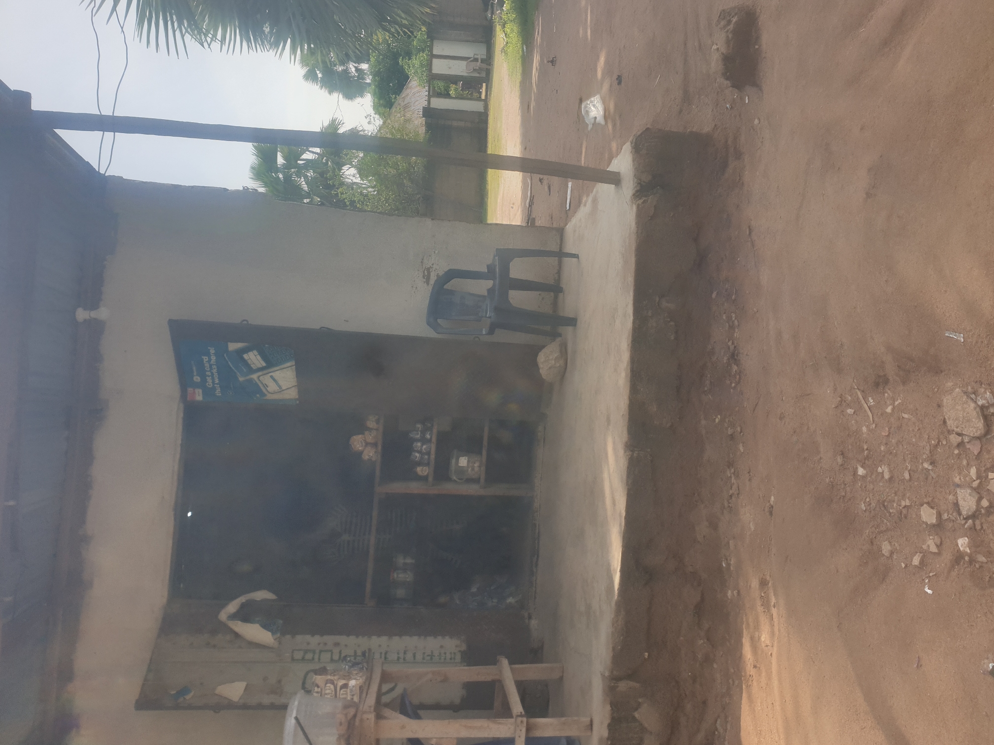 This is a photograph of a small, open-fronted shop in Jauro Yinu, Nigeria. The shop is constructed of concrete and has a corrugated metal roof. Inside the shop, shelves are stocked with various items, including jars and what appear to be food products. A wooden counter or table stands to the left of the entrance. Outside the shop, a black plastic chair is placed on the concrete porch, and a large rock sits to its left. The ground in front of the shop is dirt and appears somewhat uneven, with some scattered stones and debris. In the background, palm trees and other vegetation are visible, along with what appears to be a fence and a building with a corrugated metal roof. The sky is overcast. There are no people visible in the immediate vicinity of the shop. A blue and white sign is affixed to the wall to the right of the entrance, featuring text and graphics related to a service or product.