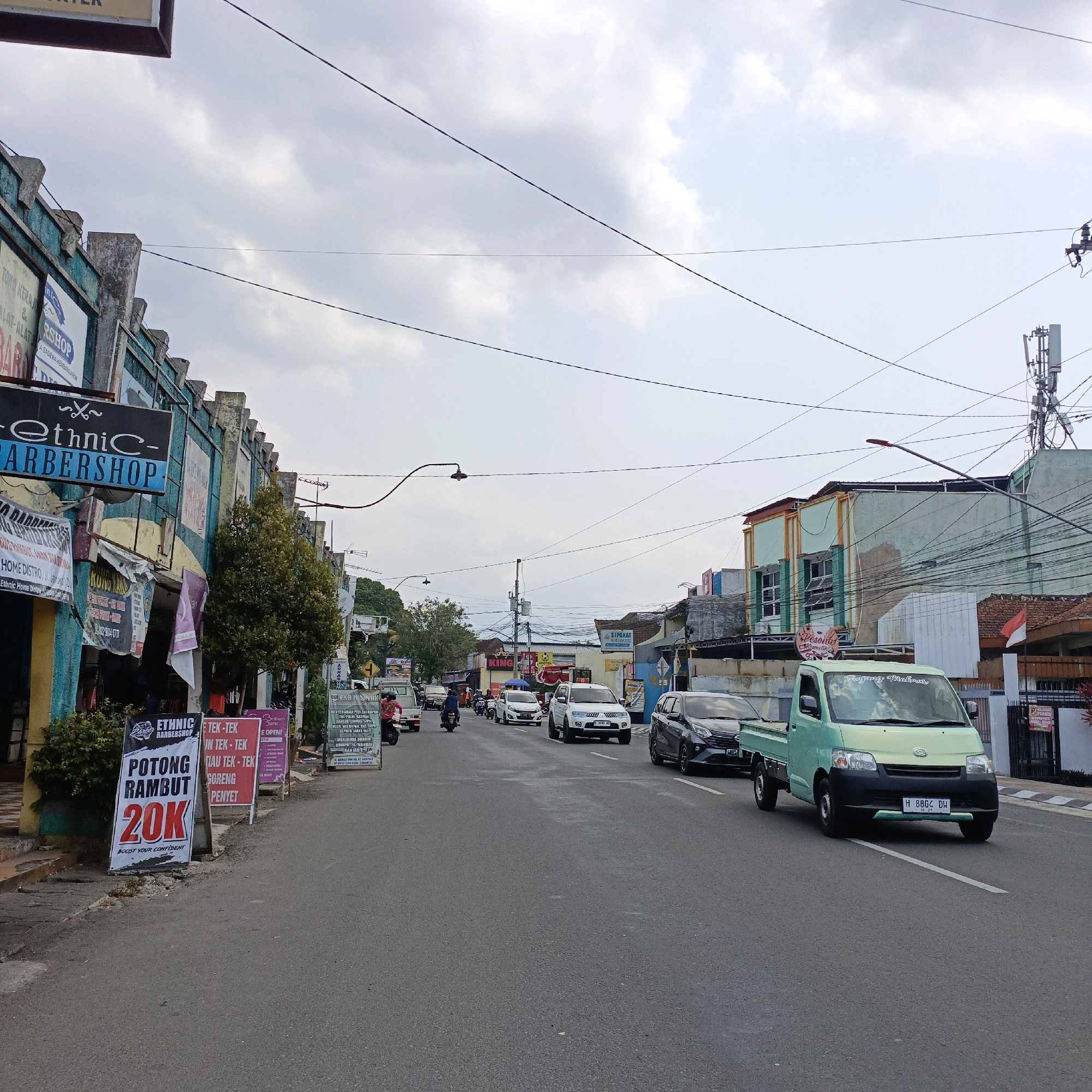 The image shows a street scene in Salatiga, Indonesia. The street is paved and there are buildings on both sides. The buildings appear to be shops, with signs in Indonesian. There are several vehicles on the street, including cars, motorcycles, and a green truck. There are also a few people visible, walking and riding motorcycles. The sky is cloudy. The overall impression is of a busy street in an Indonesian city.
Specifically, one of the signs visible is for "Ethnic Barbershop" which offers haircuts for 20,000 Indonesian Rupiah. Another sign advertises "Potong Rambut", also meaning haircut. The street has a moderate amount of traffic. The buildings are colorful, with some showing signs of age. Overhead, a complex network of electrical wires is visible, along with a telecommunications tower in the distance.