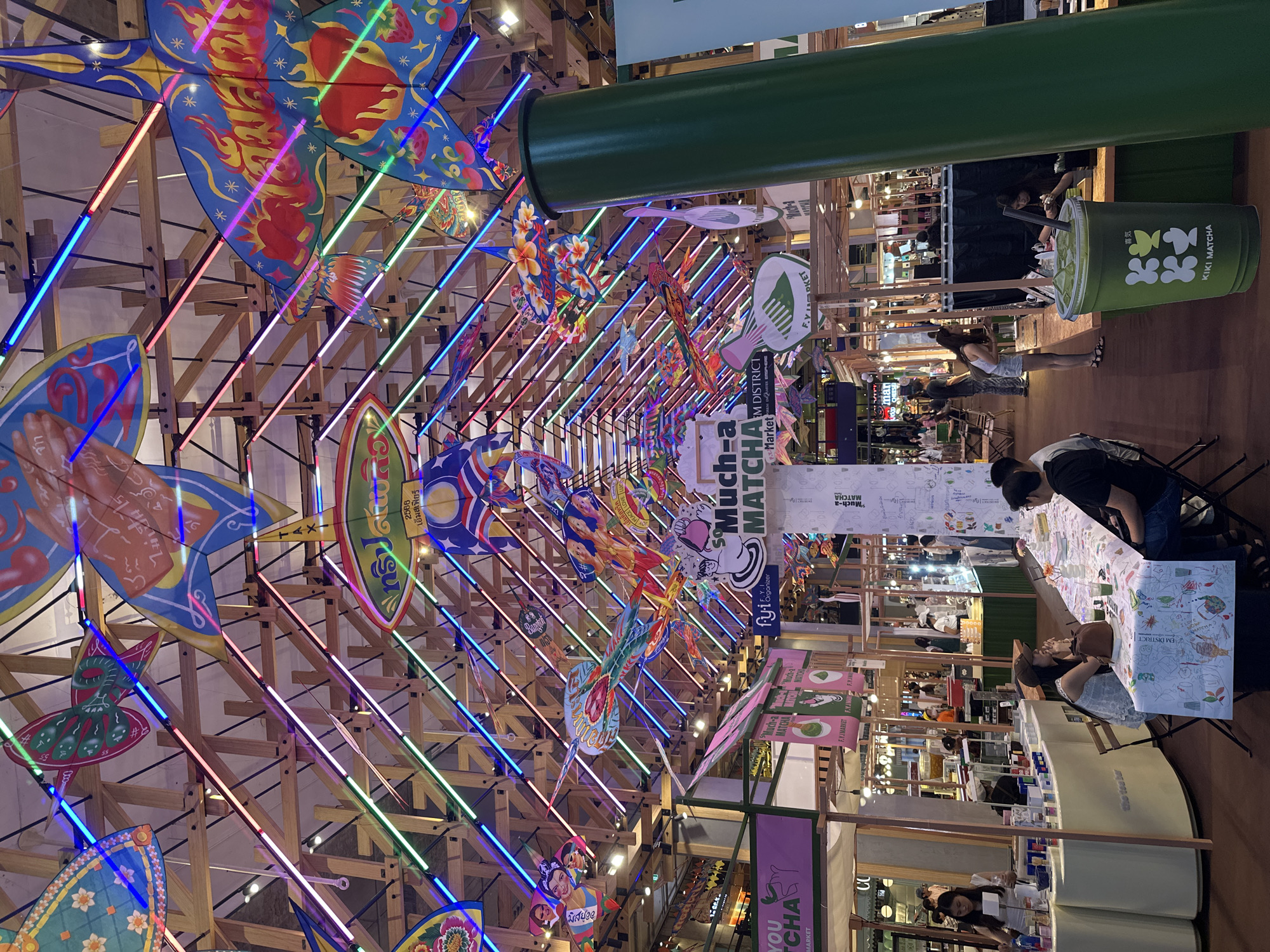 This vibrant image captures an indoor market or event space, likely within a shopping mall in Bangkok, Thailand, indicated by visible text like "EM DISTRICT" and "เอ็มสเฟียร์" (EmSphere). The scene is dominated by an elaborate ceiling decoration featuring numerous colorful kites of various shapes and sizes, suspended from a wooden beam structure. These kites are adorned with intricate patterns, including flames, flowers, hearts, strawberries, abstract designs, and even faces, with one kite explicitly featuring "Bangkok." Long, parallel neon light tubes in blue, pink, and green crisscross the wooden framework, casting a dynamic, festive glow.
Below this decorative canopy, several market stalls and booths are visible. People are casually dressed, engaged in browsing products, making purchases, or sitting at tables. A prominent, large green cup-shaped sign for "KIKI MATCHA" is visible, alongside multiple banners and signs advertising "Much-a MATCHA" and "F.Y.I. MARKET," suggesting a food and beverage, particularly matcha-themed, event or pop-up market. A long, green pipe or structural element runs horizontally across the upper mid-ground of the image. Other visible text includes "YOU CHA," "the tea bar," "2566" (Buddhist year, equivalent to 2023), "TAXI," and "LOVE." The overall atmosphere is lively, colorful, and engaging, indicative of a public event or fair.