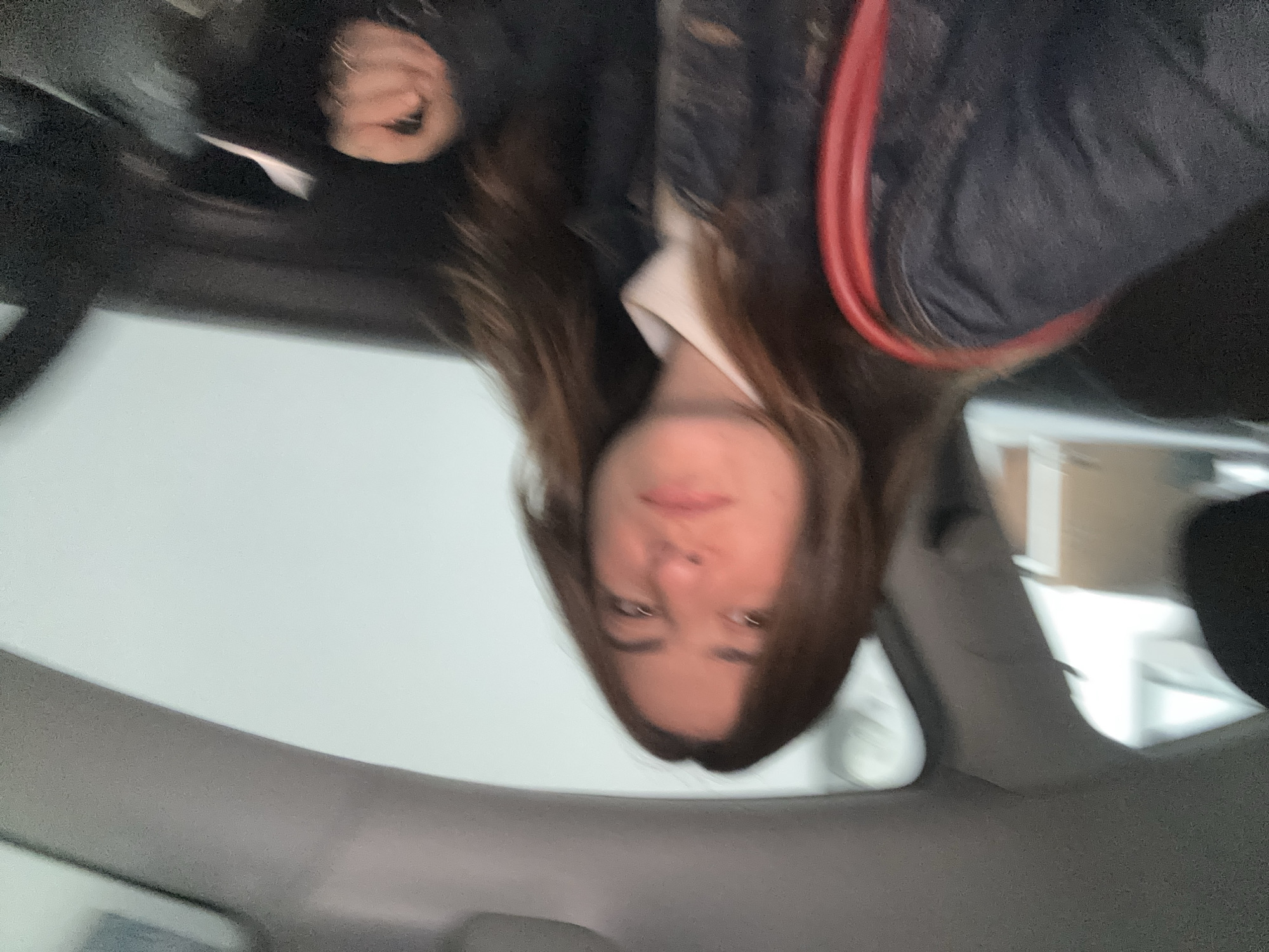 A blurry, upside-down photo taken from inside a car. The person taking the photo is in the driver's seat, as the interior of the vehicle is visible. The dashboard and roof of the car frame the bottom and sides of the image. A young woman with long, brown hair and fair skin is looking at the camera. She is wearing a white top and a dark jacket with a red strap. The location cannot be determined from the image.