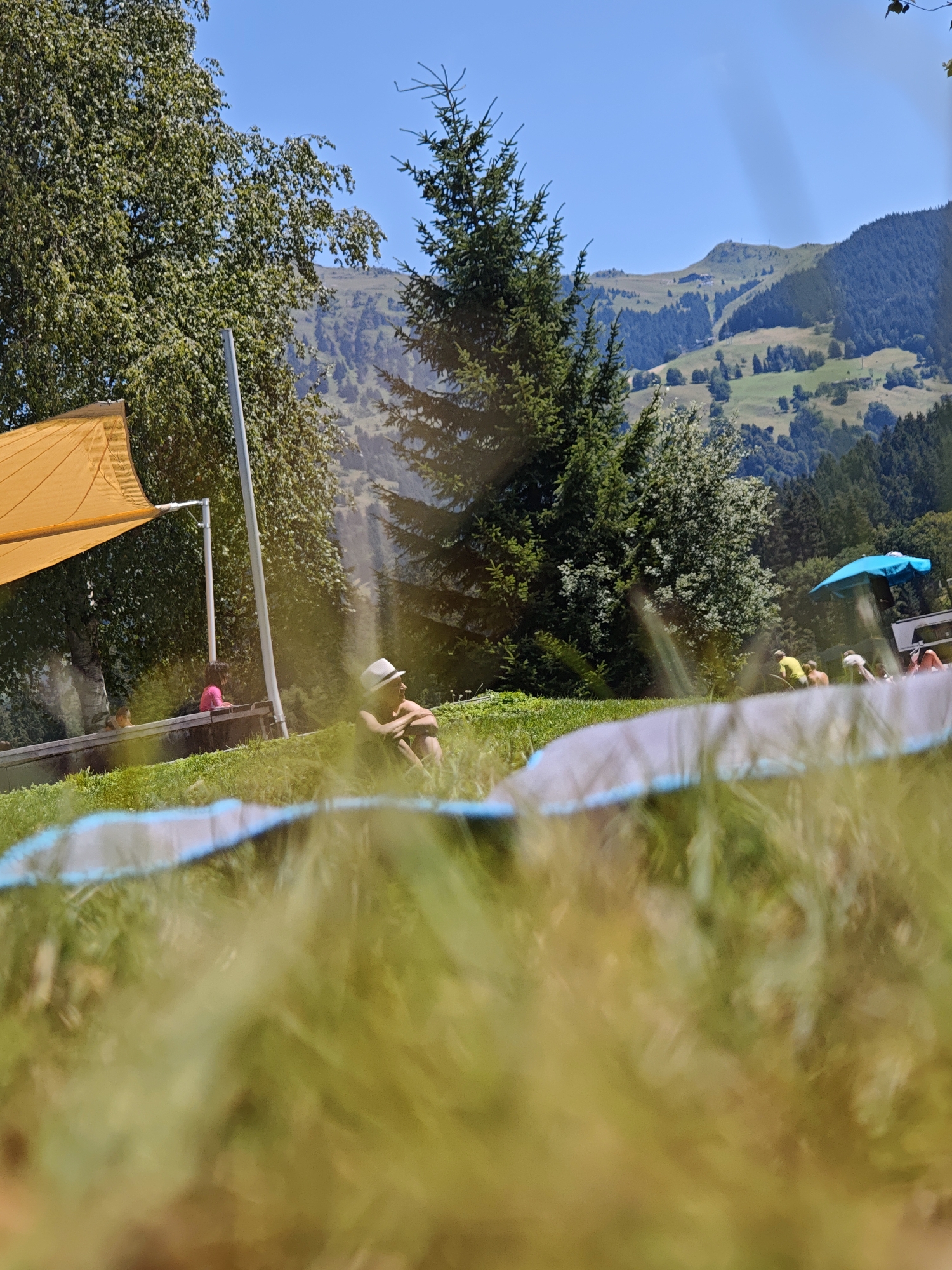 An outdoor scene, likely in Kitzbühel, Austria, captures a summer day with people relaxing in a grassy area. The foreground is dominated by blurred green grass, suggesting a low-angle shot. In the mid-ground, a person wearing a hat sits on the grass, appearing to rest. Further back, there's a group of people near a structure covered by an orange awning, with some sitting on what appears to be a low wall. Beyond them, trees and rolling hills covered in greenery and a few scattered buildings form the backdrop under a clear blue sky. A bright blue umbrella is visible on the right side of the image, near a camper van. The overall impression is one of leisure and enjoyment of a scenic mountain environment.