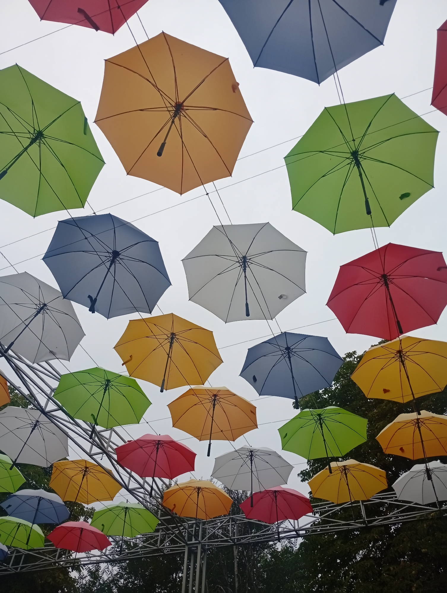 An outdoor installation features a multitude of colorful umbrellas suspended in the air against a cloudy sky. The umbrellas are various shades of red, yellow, green, and blue, and are hung at different heights and angles. Some umbrellas appear to be arranged in rows, while others are more randomly scattered. Structures supporting the umbrellas are visible in the lower part of the image, consisting of metal beams and wires. Green foliage from trees can be seen in the background, suggesting an outdoor, possibly park-like setting. The overall impression is a whimsical and decorative display.