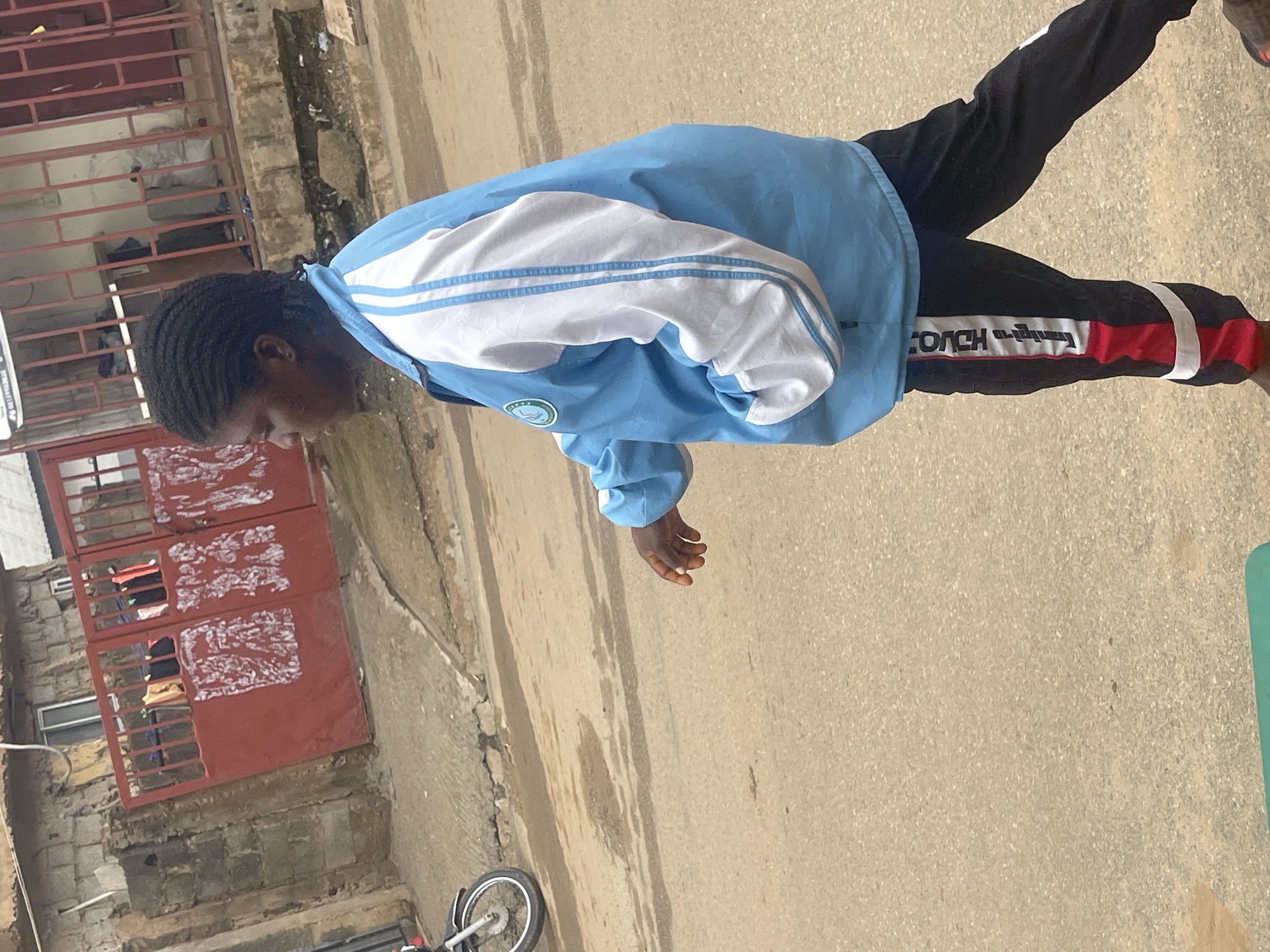 A medium shot captures a person walking on a dusty street in Abuja, Nigeria, during daylight. The individual, viewed from the side, wears a light blue jacket with white stripes on the sleeves and black pants with red and white accents. Their dark hair is braided. In the background, a building with a red gate and clothes hanging on a line is visible. A portion of a motorcycle is also present on the left side of the frame. The ground is paved with asphalt and shows signs of moisture.