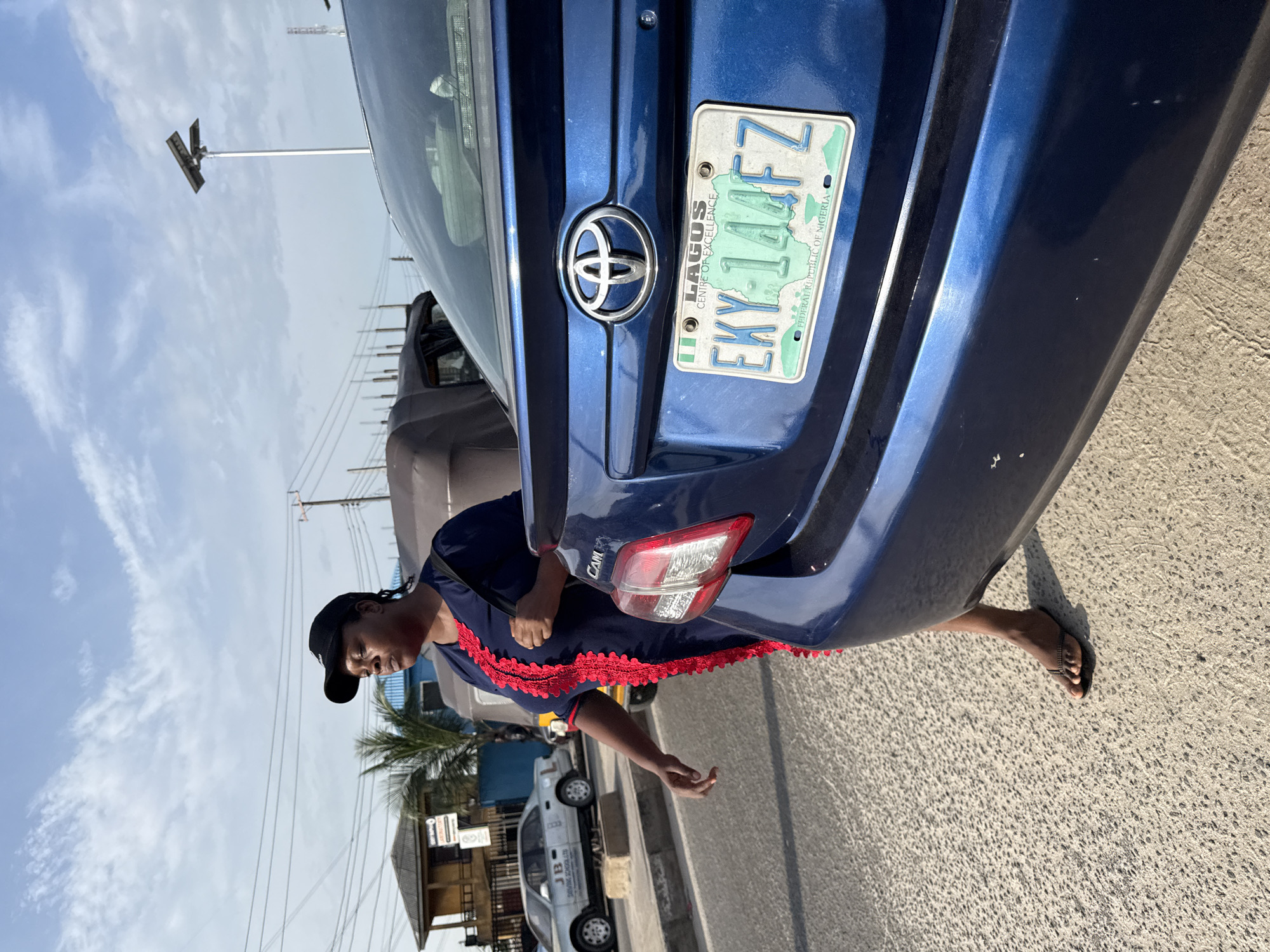 A blue Toyota car is positioned near the side of a road in an urban area in Ado, Nigeria. A woman wearing a dark blue dress with red trim, a black cap, and sandals stands next to the car's open trunk. The car's license plate reads "EKY-144FZ." There are other cars parked in the background on the other side of the street, along with various buildings and utility poles. The sky is bright with scattered clouds.