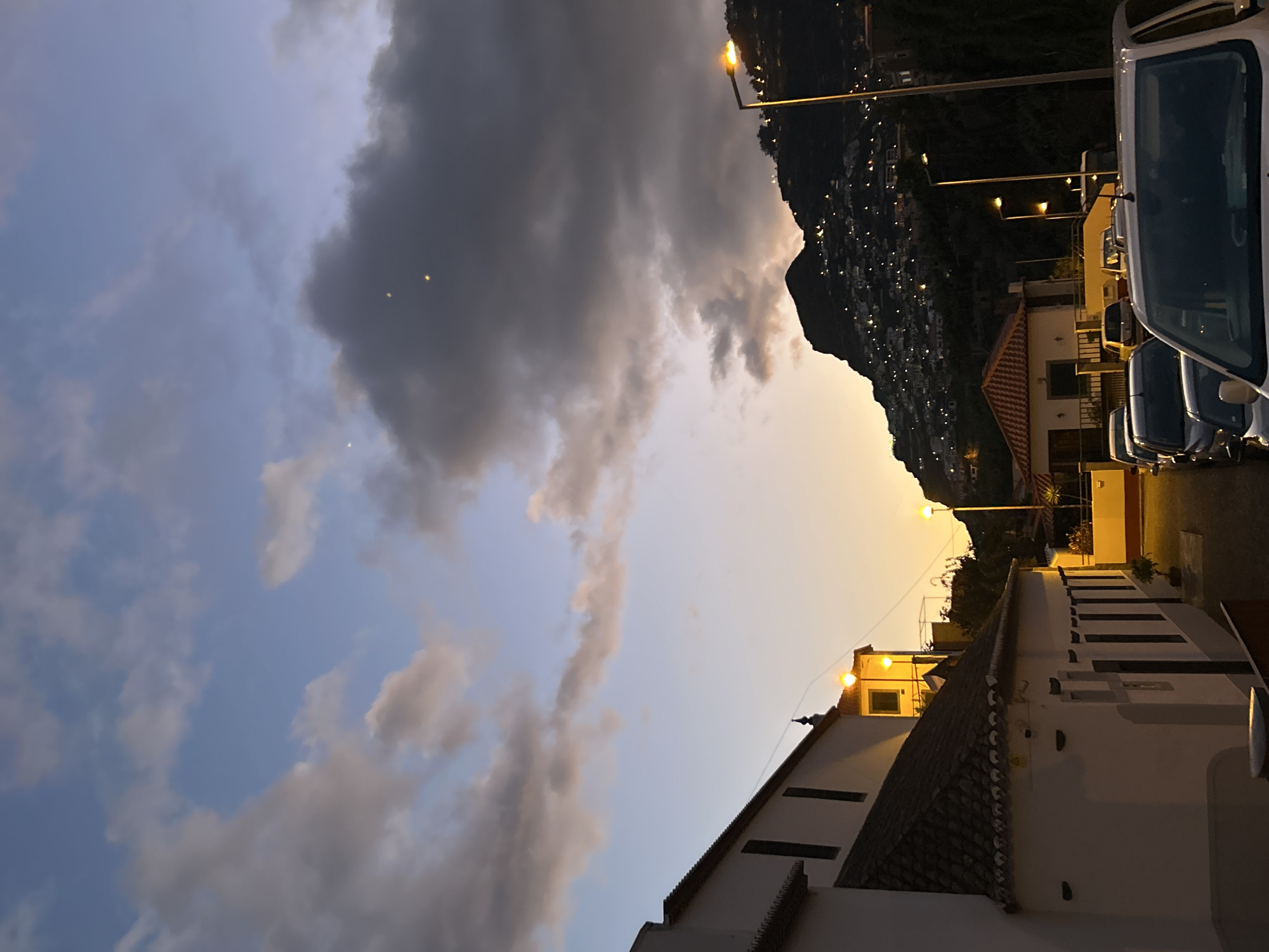 Captured from a low angle, the image shows a scene from Câmara de Lobos, Portugal, at dusk. The sky is partially obscured by a large, dark cloud formation on the left, contrasting with a gradient of blue and orange hues. Lights dot the hillside on the right, suggesting a densely populated area climbing the terrain. Street lights illuminate the scene. Buildings line the street, and cars are parked nearby.
