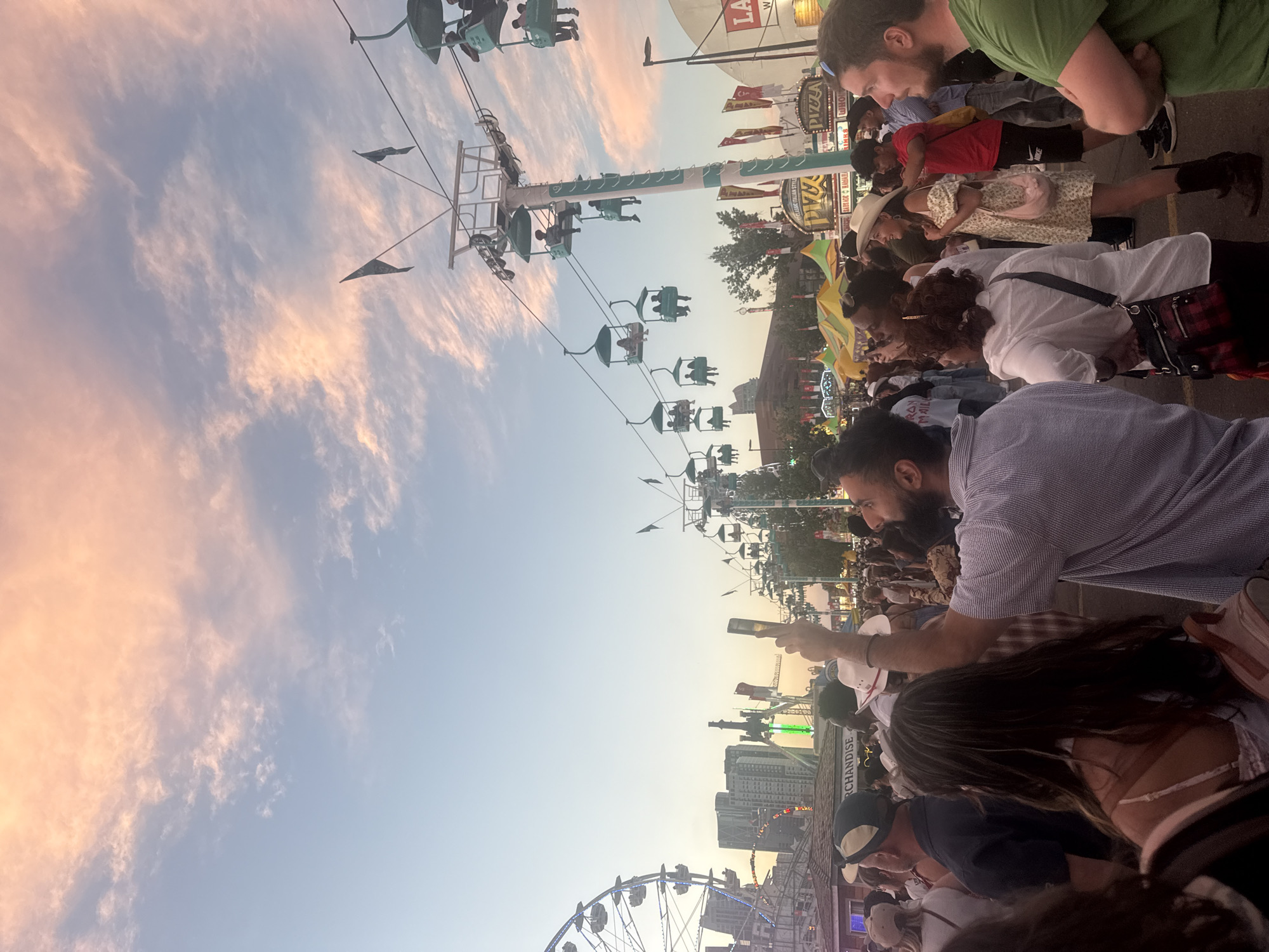 The image depicts a crowded scene at the Calgary Stampede in Calgary, Canada, at dusk. A Ferris wheel is visible on the left, and a series of gondola lifts are in operation across the middle and right of the frame. Many people are standing and looking around, some holding up their phones. The sky is filled with soft pink and orange clouds, characteristic of a sunset. Various food stalls and fairground attractions are present in the background. The overall atmosphere suggests a lively event with a significant number of attendees enjoying the attractions and the evening sky.