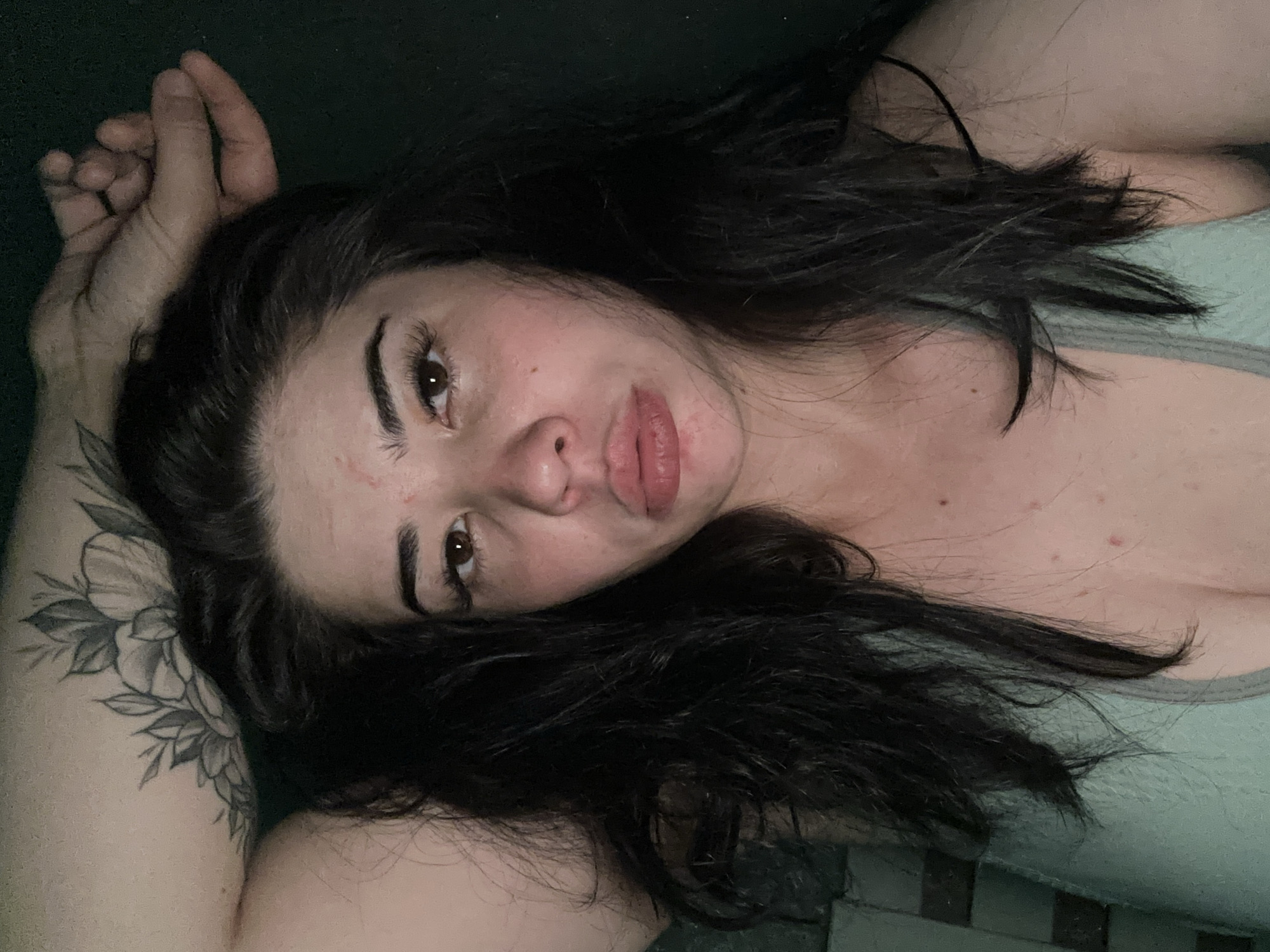 A close-up, high-angle, dimly lit, medium-shot photograph shows a young woman with long, dark, wavy hair lying down. Her head is tilted back, and her right arm is bent and resting behind her head, with her hand touching her forehead. A floral tattoo is visible on her upper right arm. She has dark brown eyes, well-defined eyebrows, and full lips. Her skin appears fair with some redness and visible pores, particularly on her cheeks and forehead. She is wearing a light blue-green tank top. The background is dark green and out of focus. The image does not provide any information about the city of Gavrilov Posad, Russia, or any other specific location.