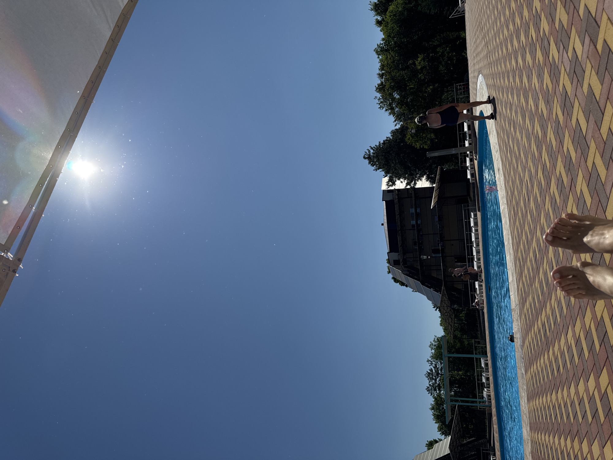 This image depicts an outdoor swimming pool area on a bright, sunny day, located in Vityazevo, Russia. The sky is a clear, deep blue, with the sun prominently visible in the upper right, generating significant lens flare and scattered light particles across the upper sky.
In the immediate foreground, the bare feet of an unseen individual are visible, resting on a patterned paved surface composed of interlocking bricks in shades of red, yellow, and brown. Beyond the feet, a large, irregularly shaped swimming pool filled with blue water stretches across the midground. To the right of the pool, a woman with gray hair, wearing a dark one-piece swimsuit, stands near the pool's edge with her back to the camera. White lounge chairs are arranged along the pool's perimeter. Other individuals are present in the mid-distance; one person is partially submerged in the pool, and others are near lounge chairs or walking in the background.
The background features several multi-story buildings, likely part of a resort or hotel complex, characterized by dark roofs and some distinct angular architectural designs. Dense green trees and foliage line the horizon behind the structures. A white fabric canopy or awning structure extends into the upper right portion of the frame, partially obscuring the sun.