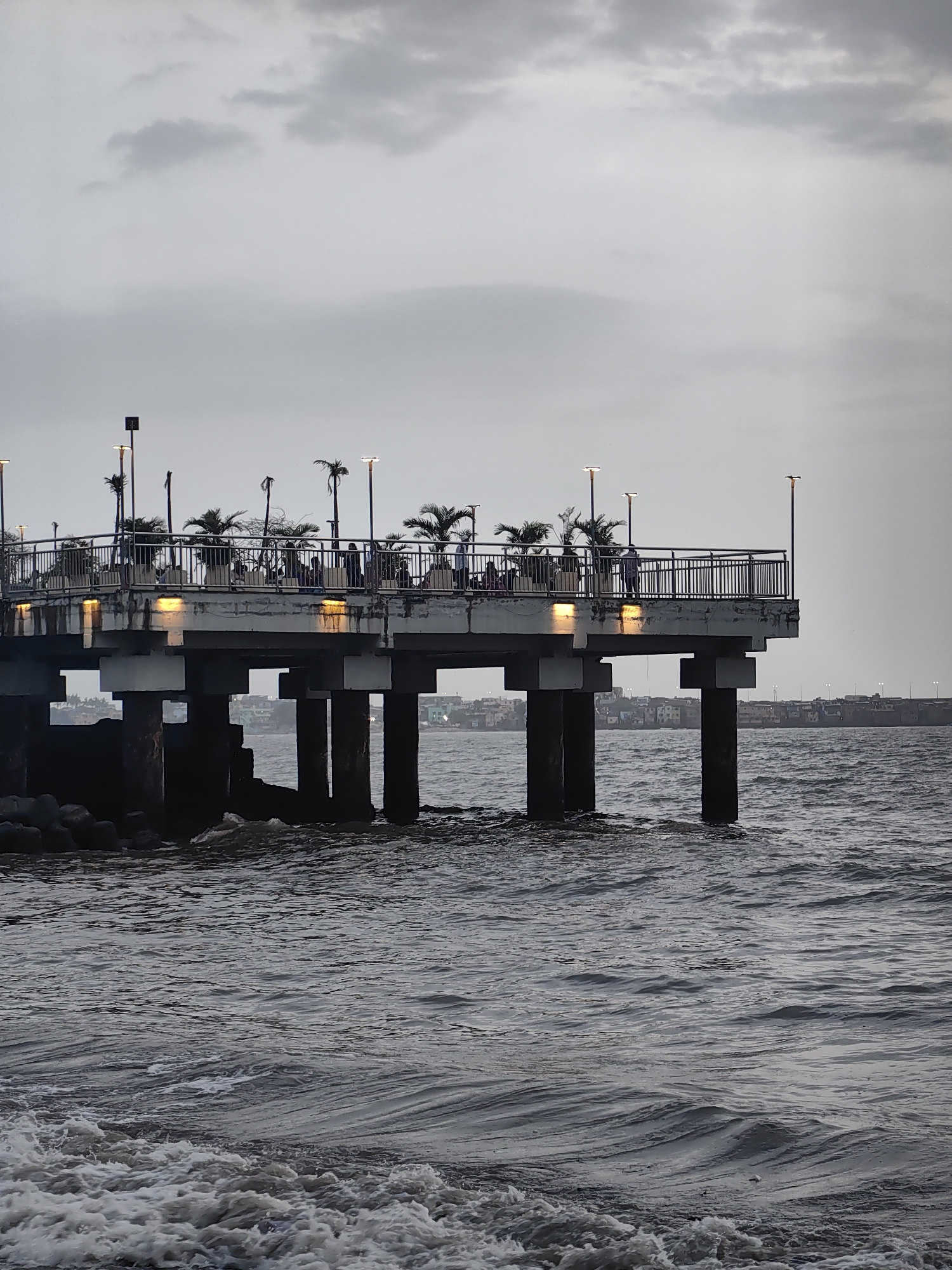 The image captures a pier extending over the Arabian Sea in Mumbai, India, under an overcast sky. The pier, constructed with concrete pillars, supports a walkway lined with railings and planters containing palm trees. Several lampposts illuminate the walkway, where a few people are visible standing and strolling. In the background, a densely populated cityscape with buildings stretches along the coastline. The sea in the foreground displays choppy waves, with foamy white crests indicating movement and depth. The overall atmosphere is subdued, likely due to the cloudy weather.