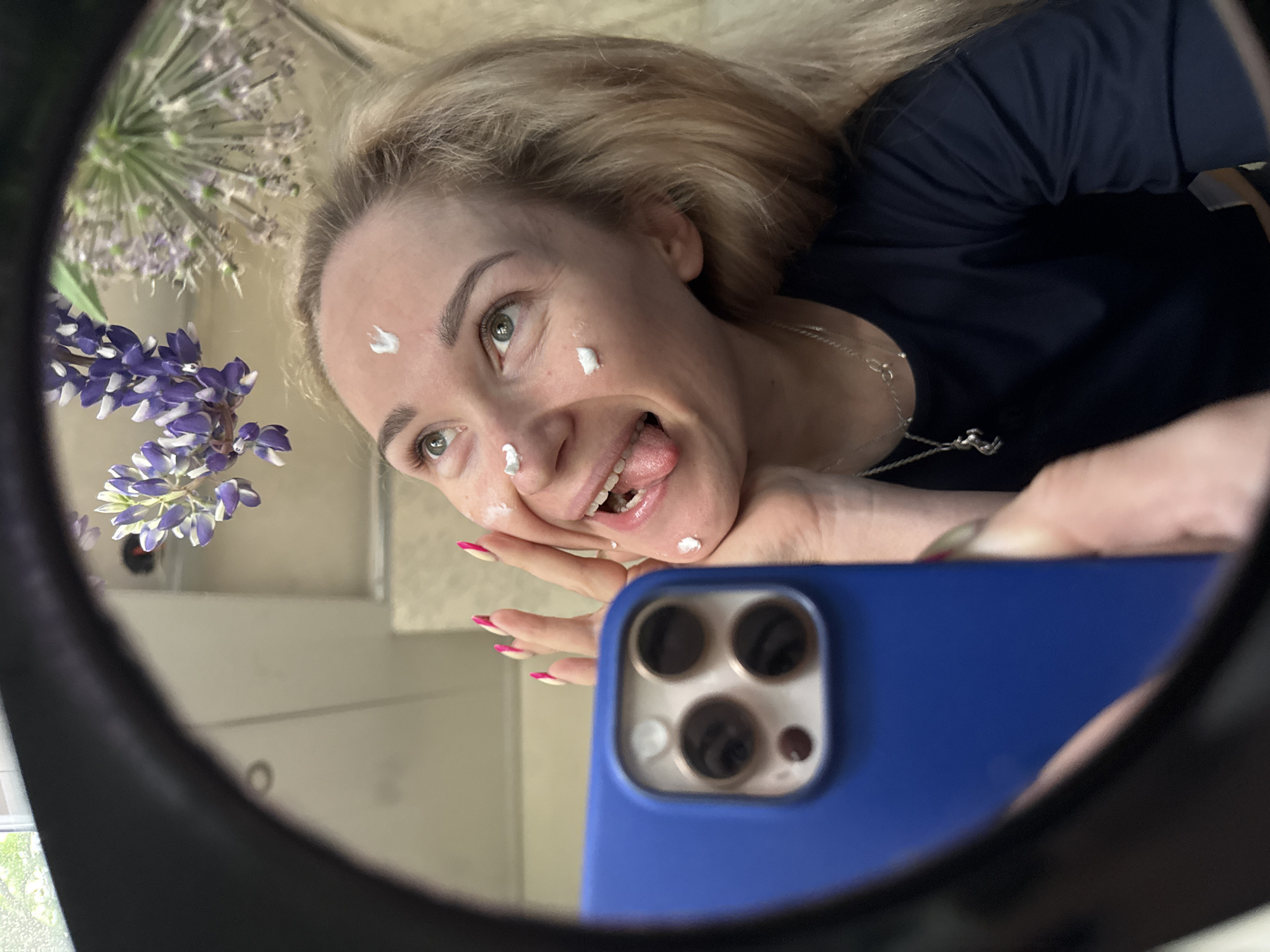 A playful mirror selfie, taken in Chaikovsky, Russia, features a blonde woman with light eyes and fair skin. Her reflection is captured in a round mirror, showing her from the chest up. She is wearing a dark blue t-shirt and a silver necklace. Several small white dots of what appears to be cream or skincare product are visible on her forehead, cheekbone, and chin.
The woman is making a cheerful and somewhat silly expression, sticking out her tongue slightly and smiling broadly. She holds a blue smartphone, identifiable by its prominent camera module, to take the picture. Her hands are visible, and her fingernails are painted with a distinct pink and white design.
In the background of the mirror's reflection, two types of flowers are present: a large, spherical purple allium and a cluster of purple lupine flowers. The presence of natural light, possibly from a window, suggests the photo was taken indoors during the daytime. The overall composition is tilted, giving the image a dynamic and informal feel. No visible text is present in the image.