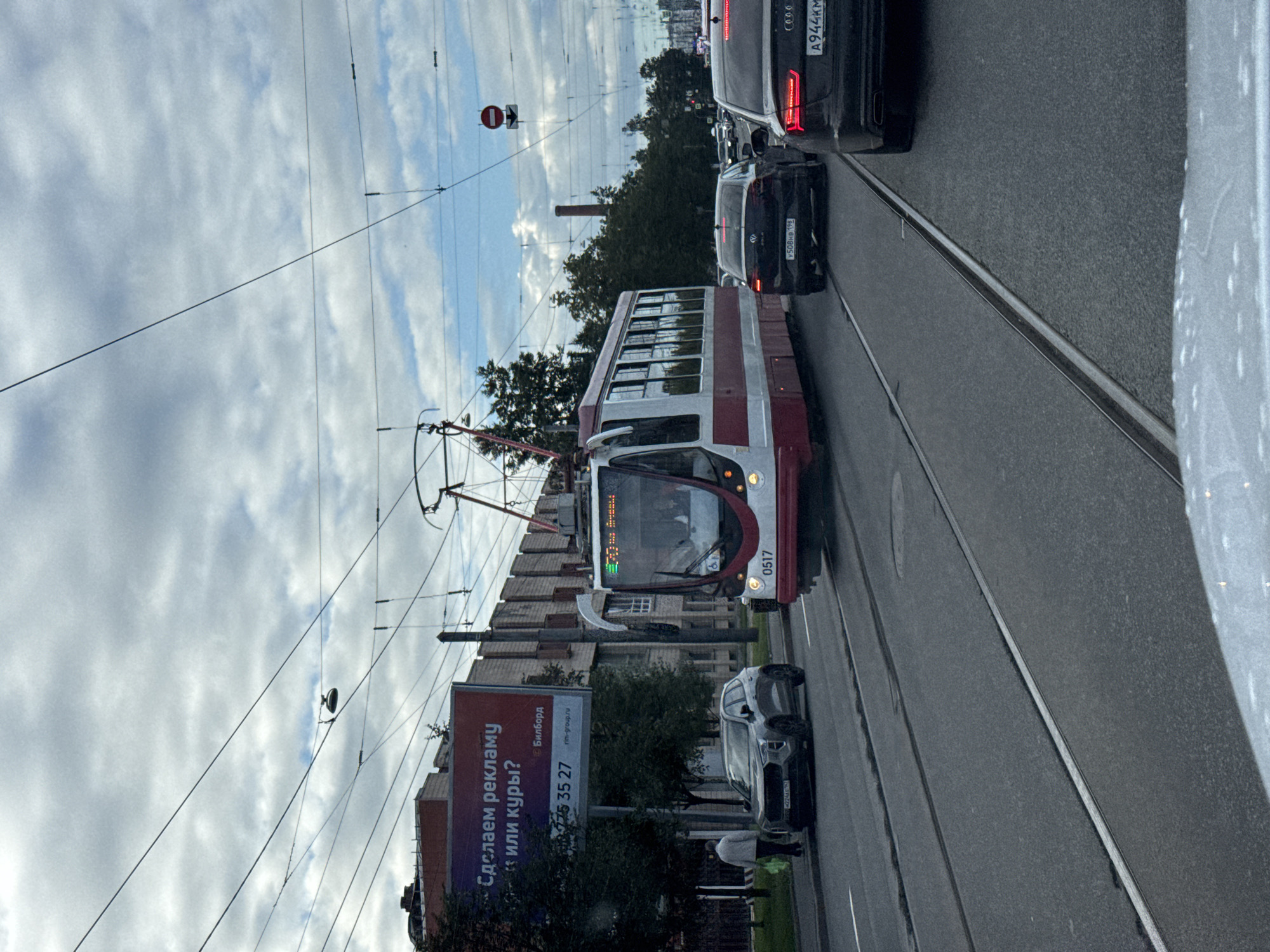 The image depicts a street scene in Saint Petersburg, Russia, on a cloudy day. A red and white tram, numbered 0517, is in motion on tram tracks. In front of the tram, a gray SUV is parked to the left, and a black Audi is in the right lane, ahead of the tram. Further down the street, another black car is visible. A billboard advertisement is on the left side of the road, with Russian text. A pedestrian, wearing a light-colored jacket, is walking on the sidewalk to the left of the SUV. Overhead wires for the tram are prominent. The environment is urban, with buildings and trees lining the street.