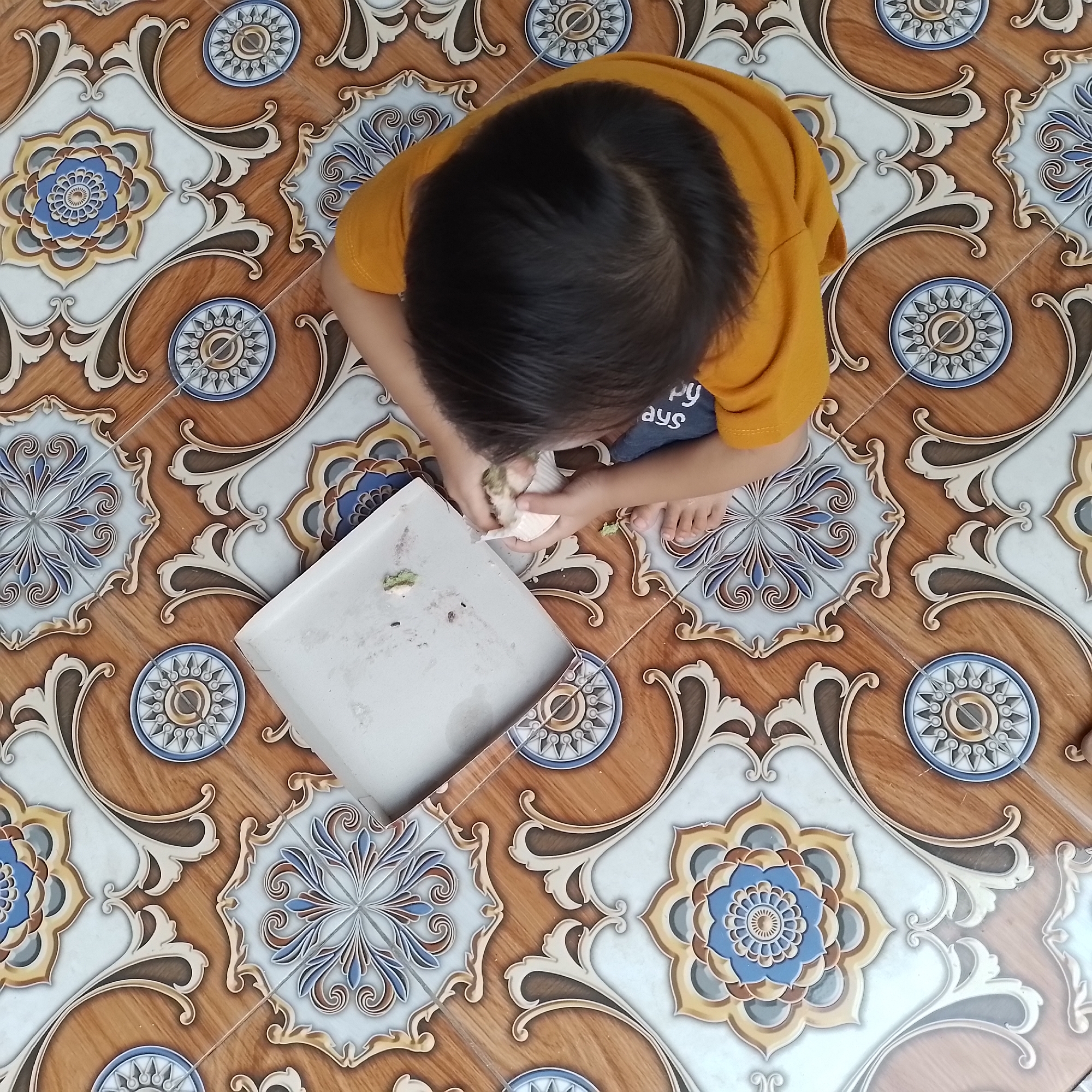 An overhead shot shows a young child sitting on a patterned tile floor in Jatisobo, Indonesia. The child, wearing a yellow shirt, is engaged with a white paper cup and a grey tray. Small green food remnants are visible on the tray and in the child's hands. The floor is decorated with intricate floral and circular motifs in shades of brown, blue, white, and gold.