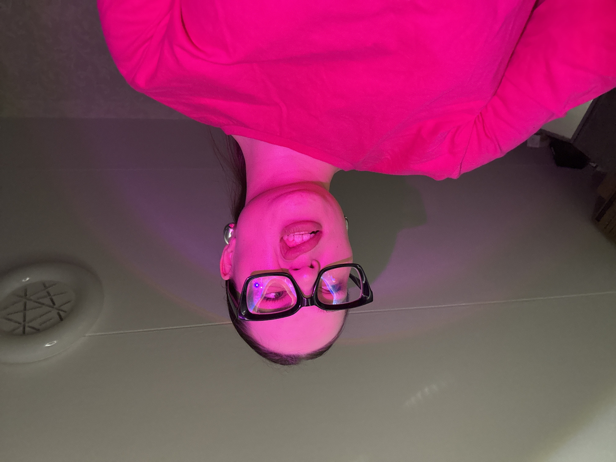 An upside-down close-up portrait shows a young individual, likely female, illuminated by vibrant pink and magenta lighting. The subject wears a bright pink long-sleeved top and distinctive black-framed glasses. Dark hair frames her face, and she looks directly at the viewer with a playful expression, her mouth slightly open with her tongue visible. The intense lighting casts a strong pink hue across her skin and clothing.
In the background, on what appears to be a light-grey ceiling or wall, a white circular ventilation-like fixture is visible in the lower left portion of the frame. A thin white line, possibly a cable or a seam, runs horizontally across the background. The area beyond these elements is dark grey, with faint indications of other objects or furniture in the distant right. A subtle, colorful light streak, resembling a small rainbow, is visible near the person's left ear. The overall impression is artistic and dynamic due to the unusual perspective and striking illumination. No text is visible in the image.