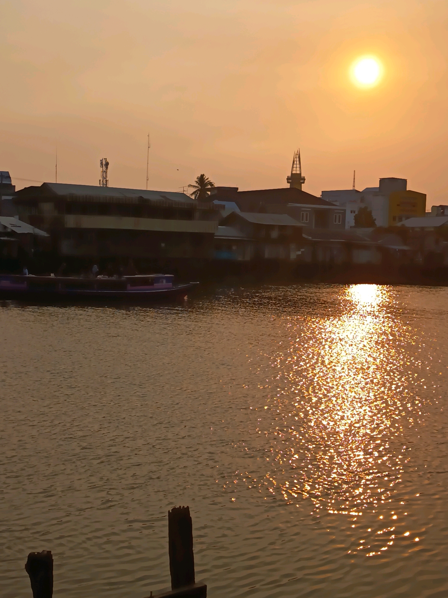 This is a horizontal, medium shot photograph taken at sunset in Banjarmasin, Indonesia. The sun, a bright orb, is visible in the upper right quadrant of the frame, casting a warm, golden glow across the scene. The sky is a soft orange hue, indicative of sunset.
In the mid-ground, a body of water, likely a river, reflects the sunlight in a shimmering pattern. A long, flat-bottomed boat, colored purple and dark blue, is docked or moving slowly on the water, with several figures visible on its deck. Along the riverbank, a series of buildings are visible. These appear to be a mix of older, single-story structures with corrugated metal roofs and some taller, more modern buildings in the background, including one with a distinct spire-like structure. Telecommunication towers are also present on the rooftops. The overall architectural style suggests a mix of traditional and contemporary urban development.
In the foreground, two wooden posts rise from the water, remnants of a pier or similar structure, adding a rustic element to the urban landscape. The water in the foreground is calmer than in the mid-ground, with subtle ripples. The lighting suggests it is late afternoon or early evening.