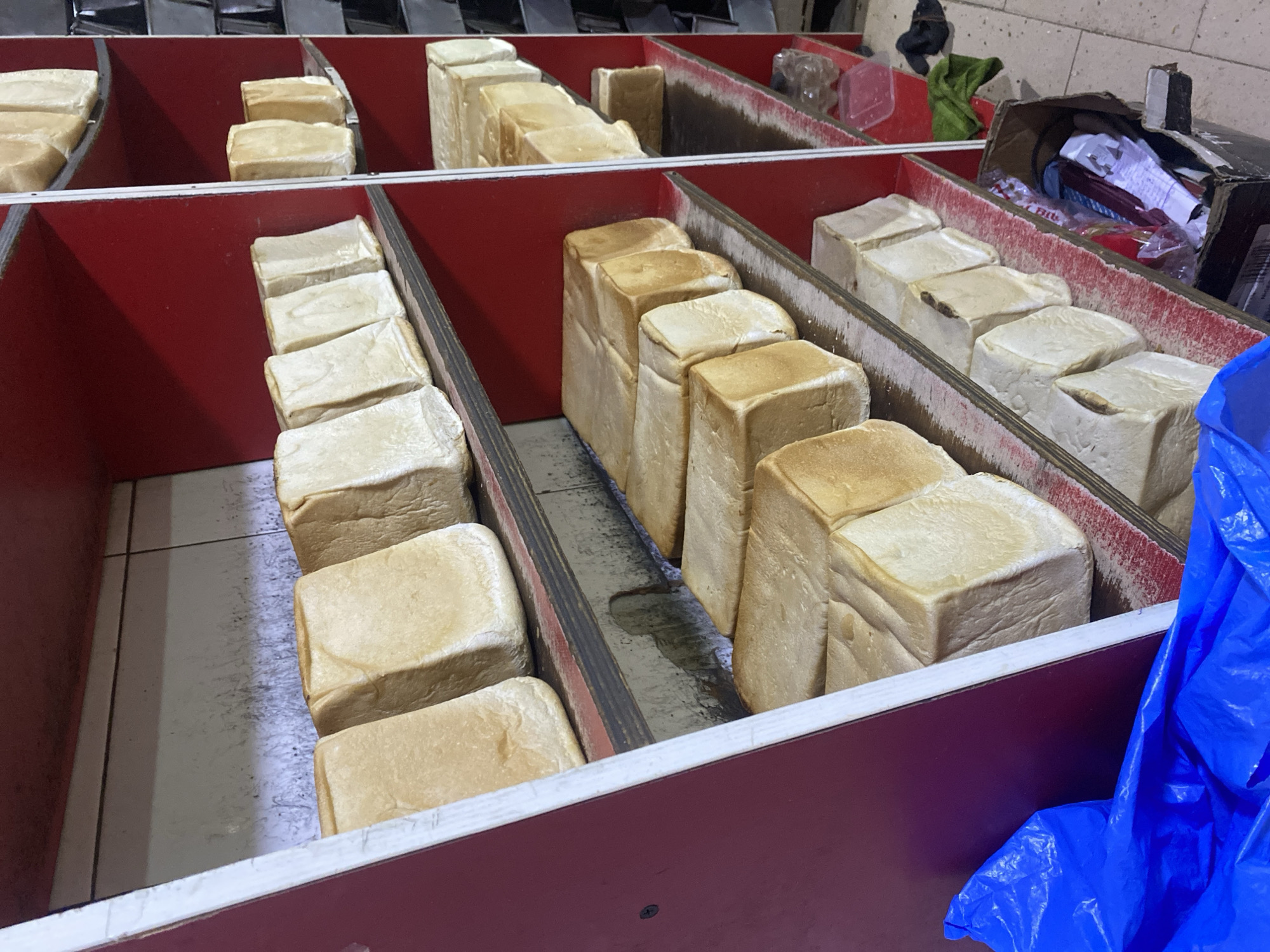 The image displays rows of rectangular loaves of bread on red-painted shelves in what appears to be a bakery or food stall in Ikorodu, Nigeria. The bread is light brown in color and has a uniform shape, suggesting it was baked in loaf pans. Some shelves are more full than others, and there are variations in the way the bread is stacked. The shelves are made of wood and appear to be somewhat worn, with some visible dust and stains. In the foreground, a large blue plastic bag is visible, partially obscuring the bottom shelf. To the right of the shelves, there are some miscellaneous items, including a plastic bottle, a pink container, and a dark piece of fabric. A cardboard box filled with papers and other items is also present on a lower shelf. The overall impression is that of a working establishment where bread is being stored and potentially sold.
