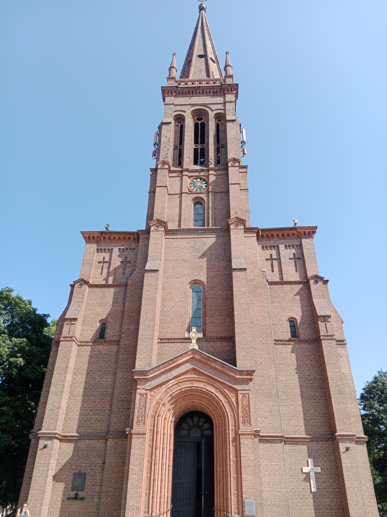 The image shows the facade of a large brick church with a tall, slender tower under a clear blue sky. The church is constructed primarily of red brick with decorative elements and arched windows. A clock is visible on the tower. Antennas are mounted on the sides of the tower. The entrance to the church is a large, arched doorway with intricate carvings. A cross is visible above the entrance and another is inscribed on the facade of the church. Trees are present on the left and right sides of the frame. A small plaque is affixed to the lower left portion of the church wall. The lower right corner of the image shows a portion of a cross inscribed on the brick wall. A person wearing a dark top and light-colored shorts is partially visible in the lower left corner of the frame, walking past the church. The inscription on the plaque is not legible.
The church depicted is the Holy Cross Church (Kościół Świętego Krzyża) located in Bydgoszcz, Poland.