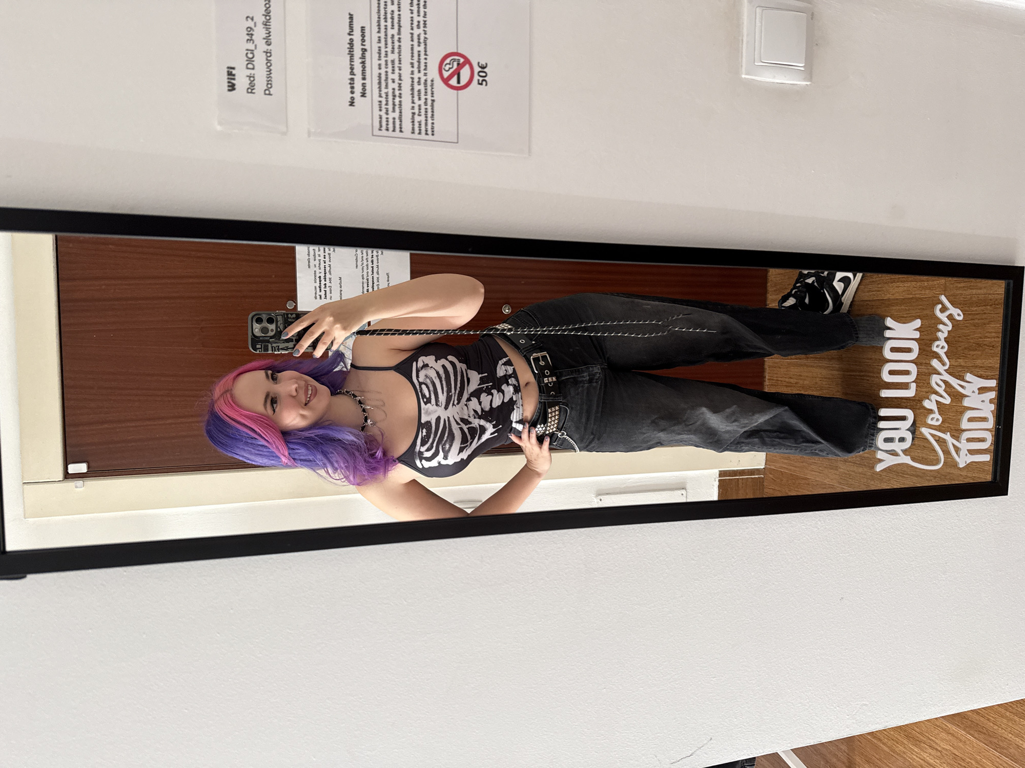 The image depicts a young woman with pink and purple hair taking a selfie in a mirror. She is wearing a black skeleton-print tank top, black jeans, and black and white sneakers. The mirror has a black frame and the phrase "YOU LOOK Gorgeous TODAY" printed on it. The mirror is hanging on a white wall, and above it, there are two signs, one about WiFi access and the other stating "No está permitido fumar" (Non-smoking room) in Spanish and English, with a 50€ penalty sign. The WIFI label indicates "Red: DIGI_349_2, Password: elwifideoz." The location, presumed to be Madrid, Spain, is inferred from the Spanish sign.