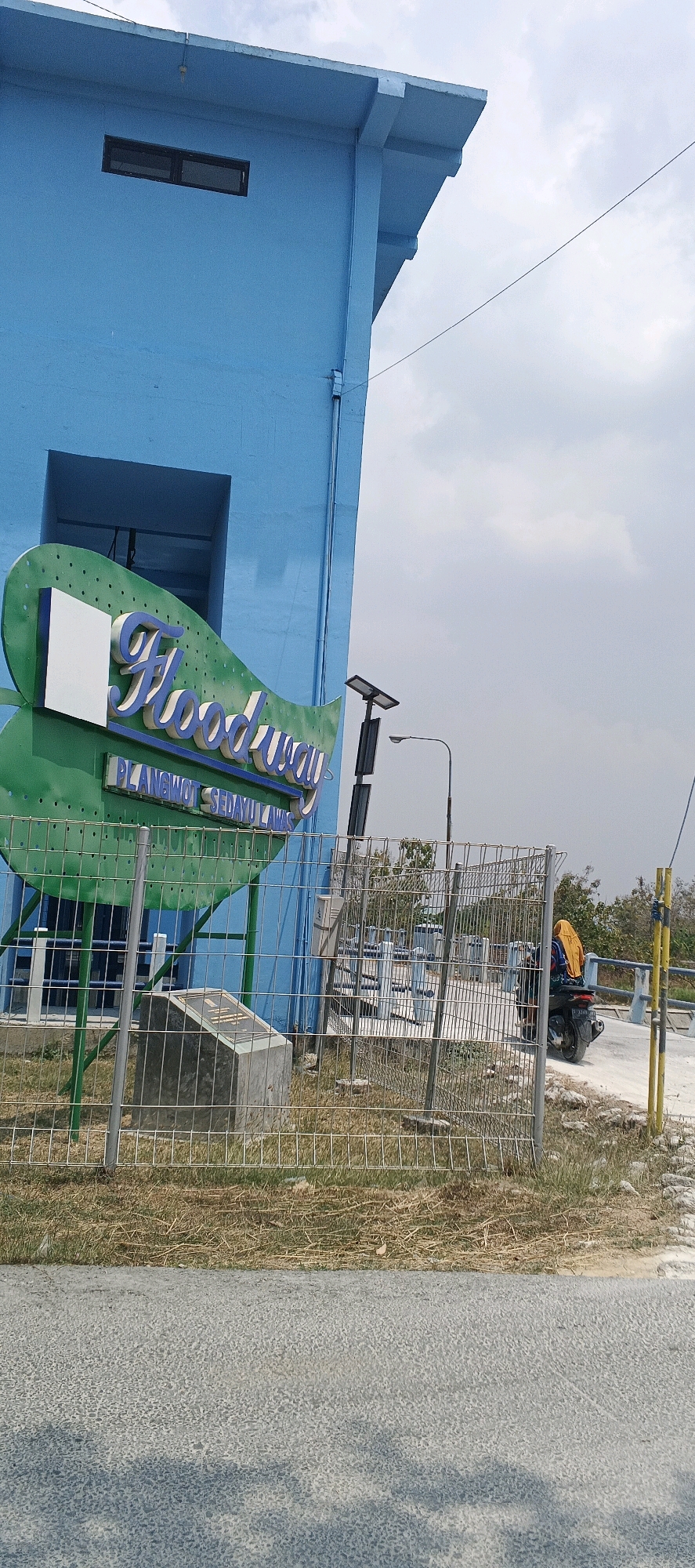 The image depicts the exterior of a blue building in Parengan, Indonesia, featuring a prominent "Floodway" sign. The sign is designed in a green, wavy shape with blue lettering, and has a white rectangular cutout on the left. Below the main sign, a secondary sign reads "PLANGWOT SEDAYU LAWAS". A chain-link fence runs across the foreground, obscuring a stone monument with a plaque on its surface. Behind the fence, various water management structures, likely sluice gates, are visible. A person wearing a yellow headscarf rides a motorcycle to the right of the frame, with a street light and solar panel assembly nearby. The environment appears dry, with sparse vegetation and a cloudy sky.
