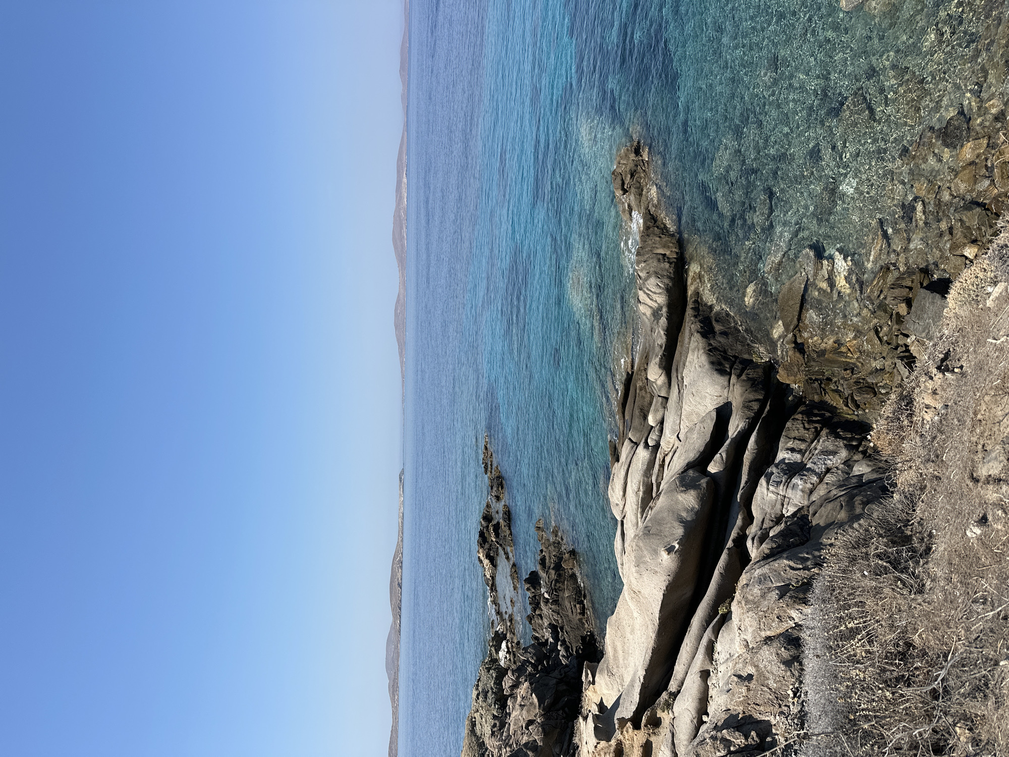 The image shows a rocky coastline on a clear day, with the bright blue sea stretching out to the horizon. In the distance, several islands are visible. The foreground features large, weathered rocks, some of which are covered in dry, sparse vegetation. The water is crystal clear, allowing the rocky seabed to be seen in shallower areas. The sky is a solid, pale blue with no clouds. The scene appears to be located in Agios Ioannis Diakoftis, Greece, a location known for its natural beauty and coastal landscapes. There are no people or specific structures visible in the image.