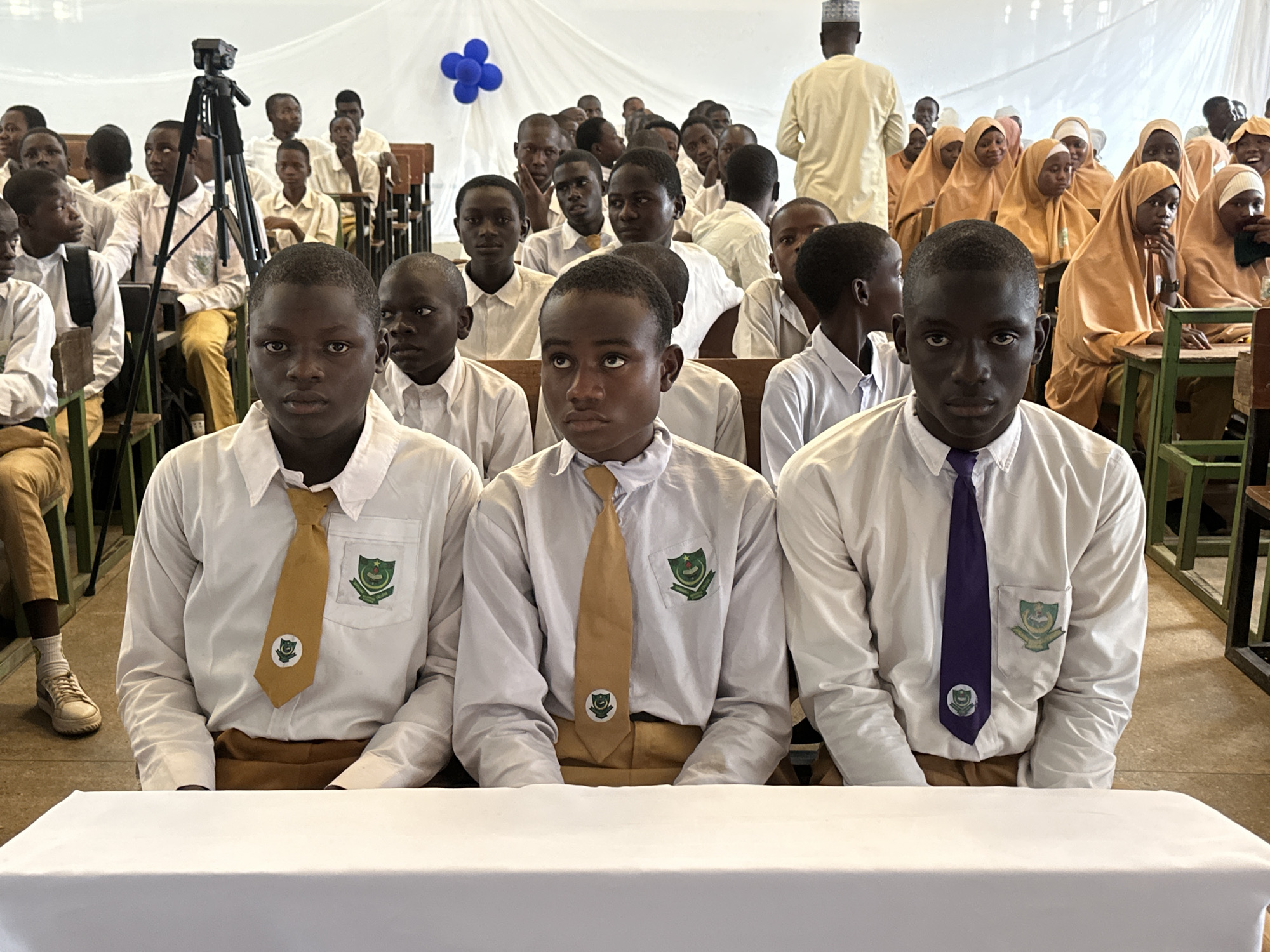 This full-frame, eye-level indoor shot captures a formal gathering of students at a school in Unguwan Dosa, Nigeria. In the immediate foreground, three young male students are seated behind a white-draped table. They wear white long-sleeved shirts, light brown trousers, and ties; the two on the left have golden-yellow ties, while the one on the right has a purple tie. Each student's shirt pocket and tie display a green and white school crest, featuring a star, crescent, and other symbols within a shield. Their expressions are serious and attentive.
Behind the foreground students, other male students in similar uniforms are seated, observing the event. To the right and further back, a group of female students is visible, uniformly dressed in light orange or tan headscarves and matching garments. In the far background, a man in traditional light-colored attire and a cap stands with his back to the camera. On the left, a camera on a tripod suggests the event is being recorded. Blue balloons are clustered decoratively on the white fabric draped across the background wall, indicating a special occasion. The scene suggests an important school assembly, competition, or formal educational event taking place during the daytime.