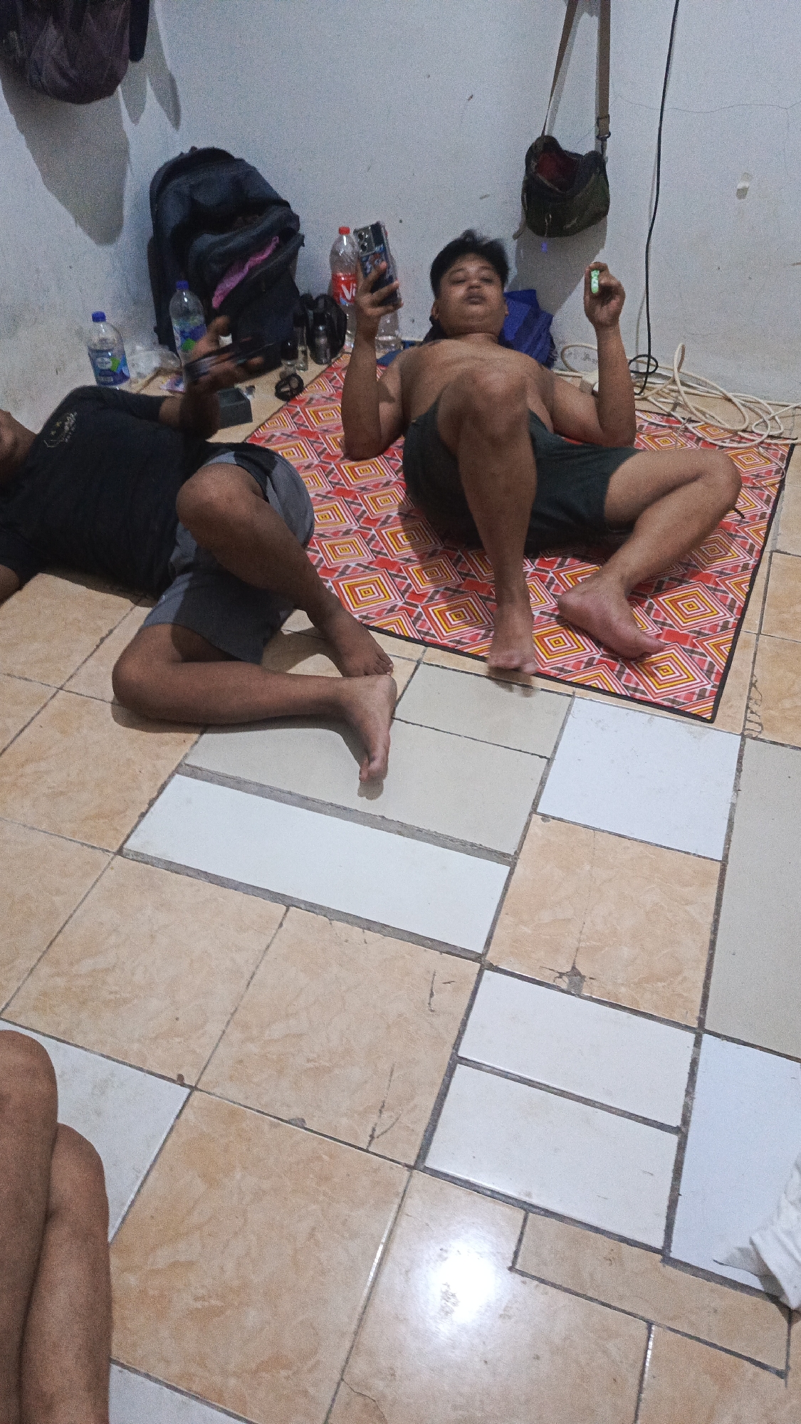 Two individuals are seen lying on a mat on the floor of a room. The room has tiled flooring in earth tones and white. The wall appears to be a light, off-white color.
One individual, positioned on the right side of the frame, is shirtless and wearing dark shorts. They are lying on their back with their legs spread. This individual is holding a smartphone in their right hand and what appears to be a glowing object, possibly a cigarette or vape, in their left hand.
The second individual is on the left side of the frame, wearing a black t-shirt and shorts. They are also lying on their back, with their legs bent. This individual is holding a smartphone.
In the background, there are several objects including backpacks, water bottles, and a bag hanging from the wall. Electrical cords are visible on the floor. The overall lighting suggests an indoor environment, possibly at night. The scene does not provide specific visual cues to pinpoint the exact location within Tangerang, Indonesia, beyond it being an indoor residential or lodging space.