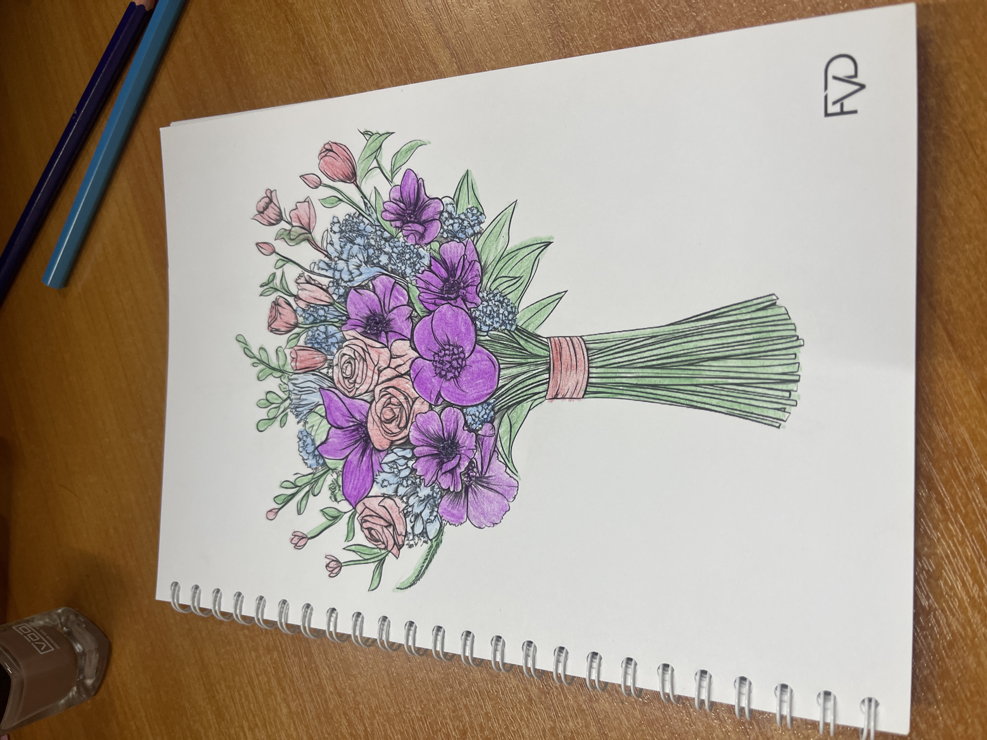 The image shows a close-up, high-angle shot of a notebook with a drawing of a floral bouquet. The bouquet is rendered in colored pencil with black outlines. It features a variety of flowers in shades of purple, pink, and light blue, with green leaves and stems. The bouquet is tied with a coral-colored ribbon. The drawing is on the right side of the notebook page, with the stems extending towards the right edge. The notebook is spiral-bound with a white page and visible metal spiral binding along the bottom edge. To the left of the notebook, there are two colored pencils, one dark blue and one turquoise, lying on a wooden surface. In the bottom left corner, a small, clear object with a label that appears to read "VGG" is visible. At the top right corner of the notebook page, there is a stylized logo or initials that read "FVD". The lighting suggests an indoor setting, possibly a desk or table, with natural light. The overall mood is calm and artistic.