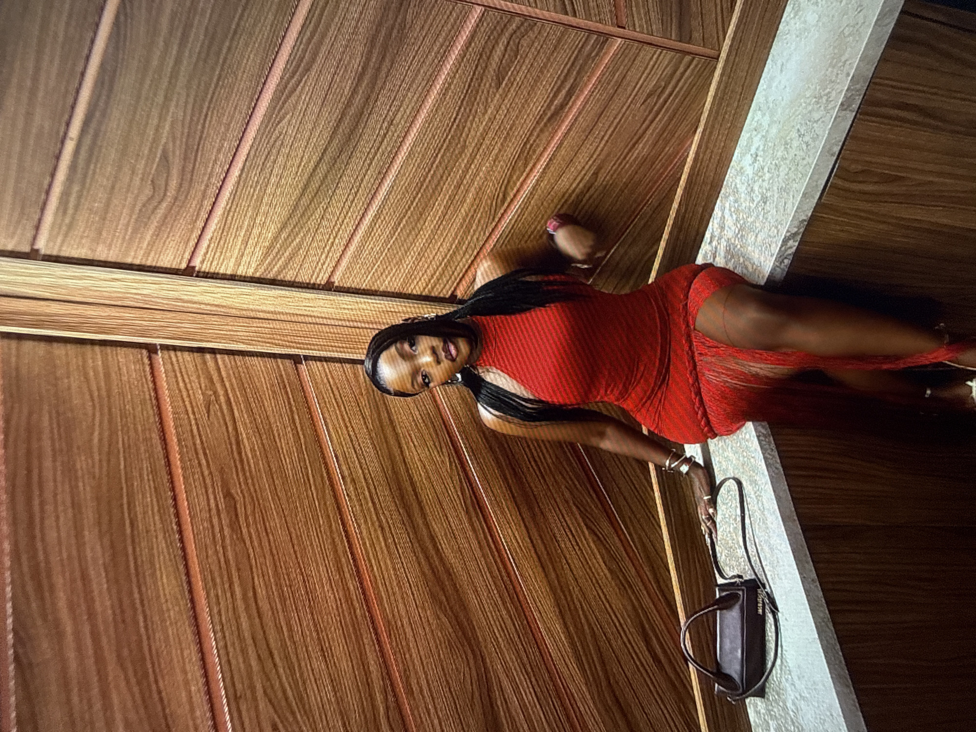 A Black woman with dark skin and braided hair wears a form-fitting, sleeveless red dress with fringe detail at the hem. She is seated on a marble counter in what appears to be an elevator, given the wooden paneling of the walls. A dark brown handbag rests on the counter to her left. She wears silver jewelry on her wrists and fingers, and a watch on her left wrist. The background is a warm-toned wooden paneling, arranged in horizontal slats. The lighting is warm, casting shadows on the woman and the environment. The location is identified as an elevator in Abuja, Nigeria.