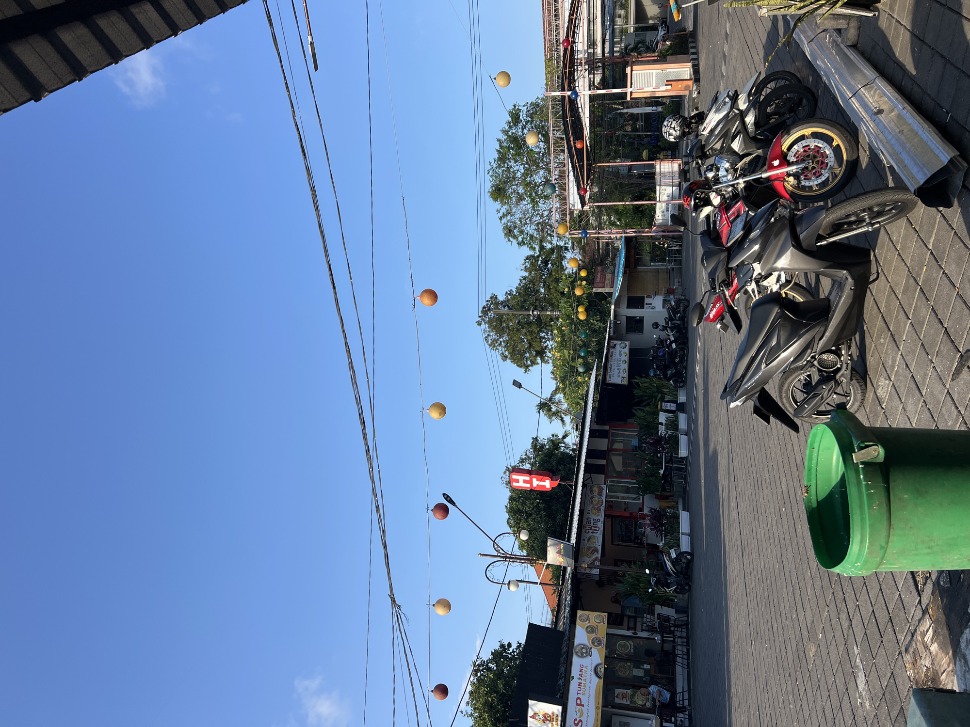 In Kuta, Indonesia, a bright, clear sky dominates the upper portion of the image, with power lines strung across it. Below, a row of commercial establishments, likely shops or restaurants, line a paved area. Colorful decorative orbs hang suspended from wires above the buildings. Several motorbikes are parked in front of these businesses, with some riders or patrons visible. A prominent red "HI" sign is mounted on one of the buildings. In the foreground, a large green trash can stands on the paved surface, and a metal barrier lies on its side. The scene suggests a bustling commercial district in an outdoor setting.