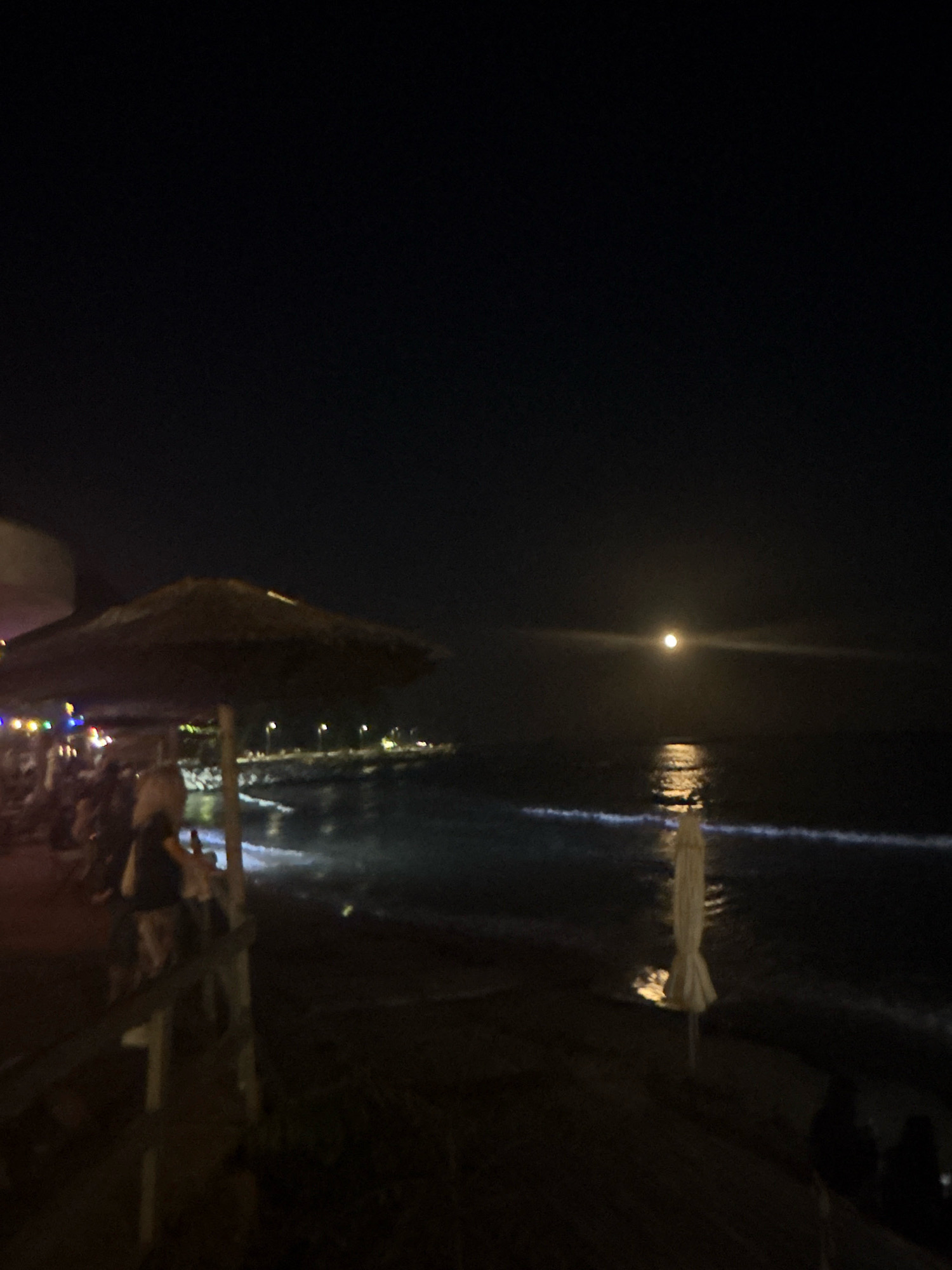 This is a nighttime, full-body, eye-level, wide-angle shot of a beach scene. In the foreground, a large straw umbrella is partially visible on the left, with a wooden railing and what appears to be a table and chairs underneath. Several indistinct figures are seated or standing near the umbrella. A second, closed, white umbrella stands in the mid-ground on the sand. The sea is in the background, with gentle waves breaking on the shore. A pier or jetty with lights can be seen in the distance on the left. The moon is high in the dark sky, casting a reflection on the water. The overall scene depicts a tranquil evening at a beachside establishment.