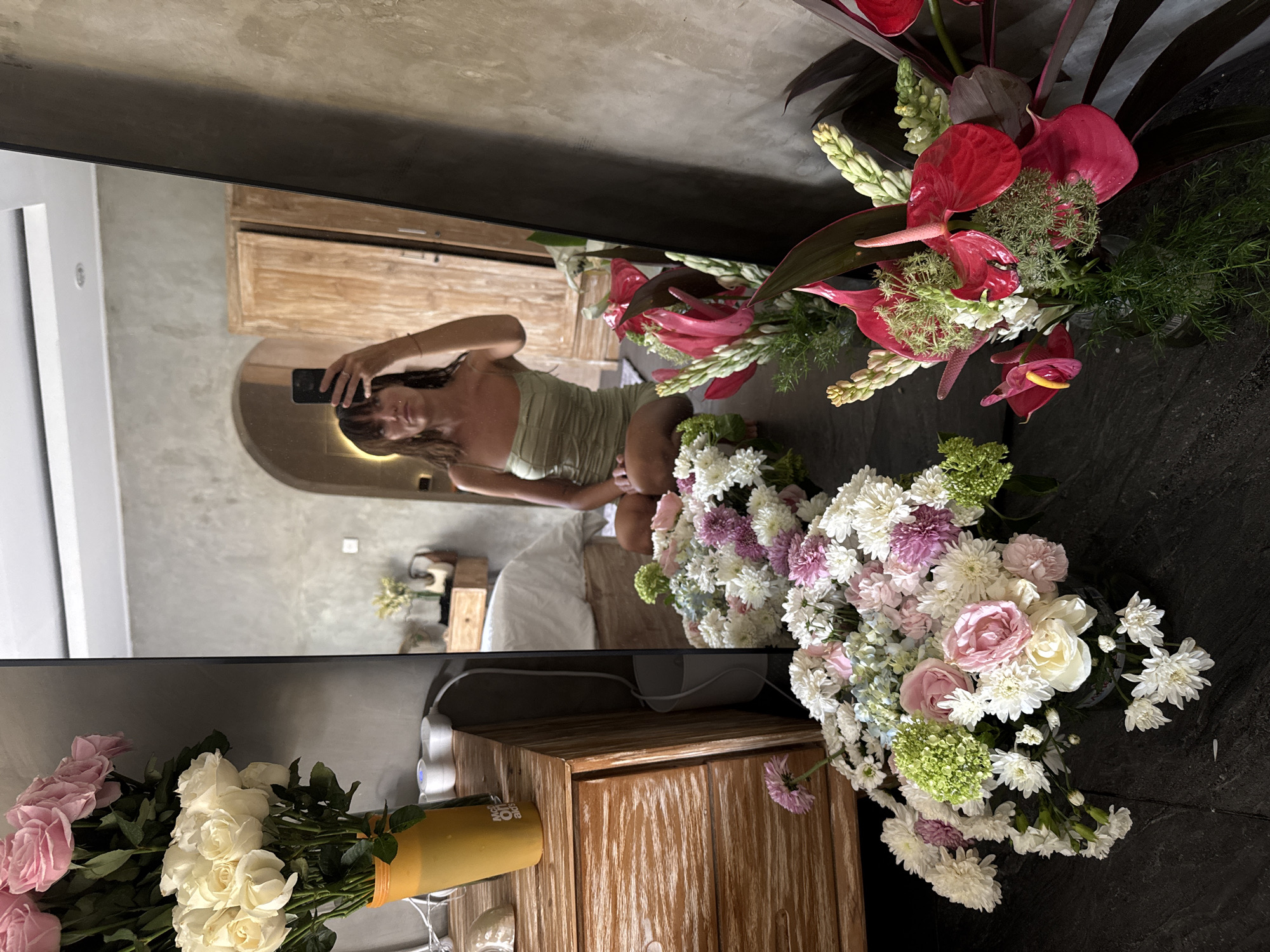 The image appears to be a selfie taken in a hotel or resort in North Kuta, Indonesia. The reflection shows a woman in a light green dress holding a phone, presumably taking the picture. The background reveals a bedroom with a bed and wooden furniture. The foreground features two large floral arrangements; one with red anthuriums and greenery, the other with mixed pastel-colored roses and chrysanthemums. A wooden cabinet is visible below the mirror. The setting suggests a high-end lodging establishment.