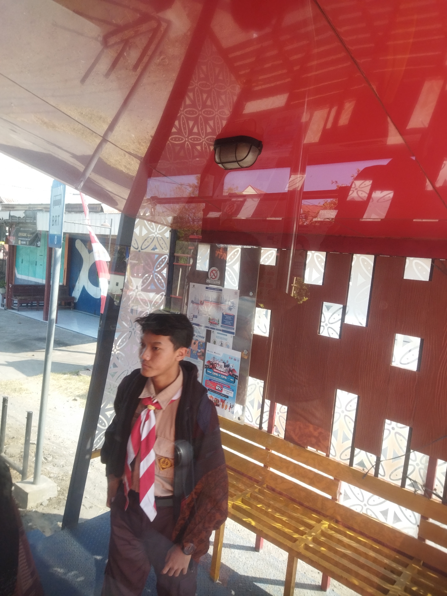 A teenage male, dressed in a scout uniform including a tan shirt, brown pants, and a red and white striped scarf, stands at a bus stop in Nguter, Indonesia. He is facing right, looking out of frame. The bus stop has a red roof and a wooden bench. The background shows a street with buildings and a sign that says "BRT." The overall scene is brightly lit by the sun.