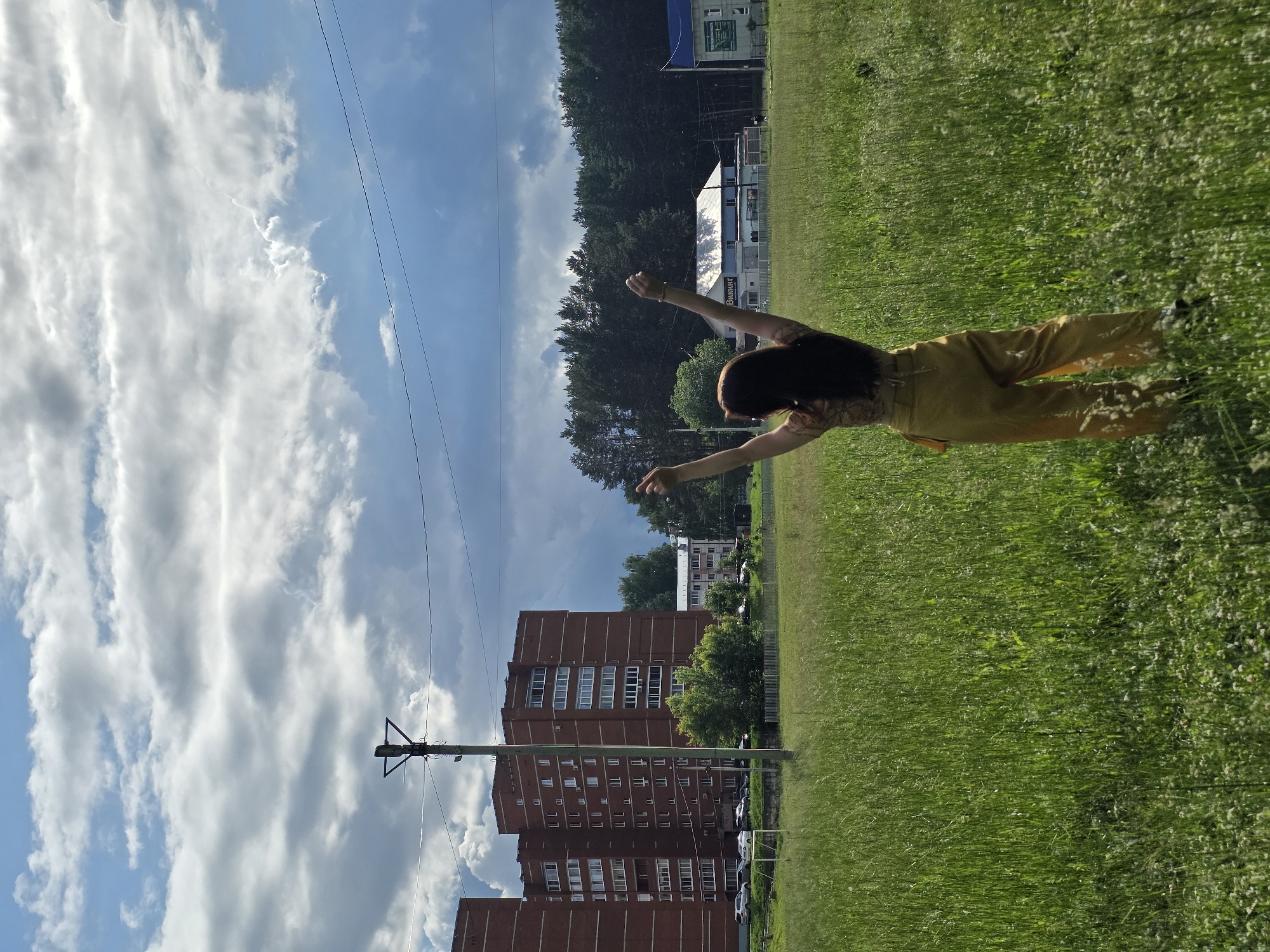 In the city of Chaikovsky, Russia, a woman with long, dark hair stands facing away from the camera in a grassy field. She is wearing a patterned top and yellow pants. Her arms are raised in the air. In the background, a tall brick apartment building is visible, along with a line of trees and other buildings. The sky is cloudy, with patches of blue. A utility pole with wires is situated to the left of the woman. The overall scene depicts an outdoor environment with urban elements present.