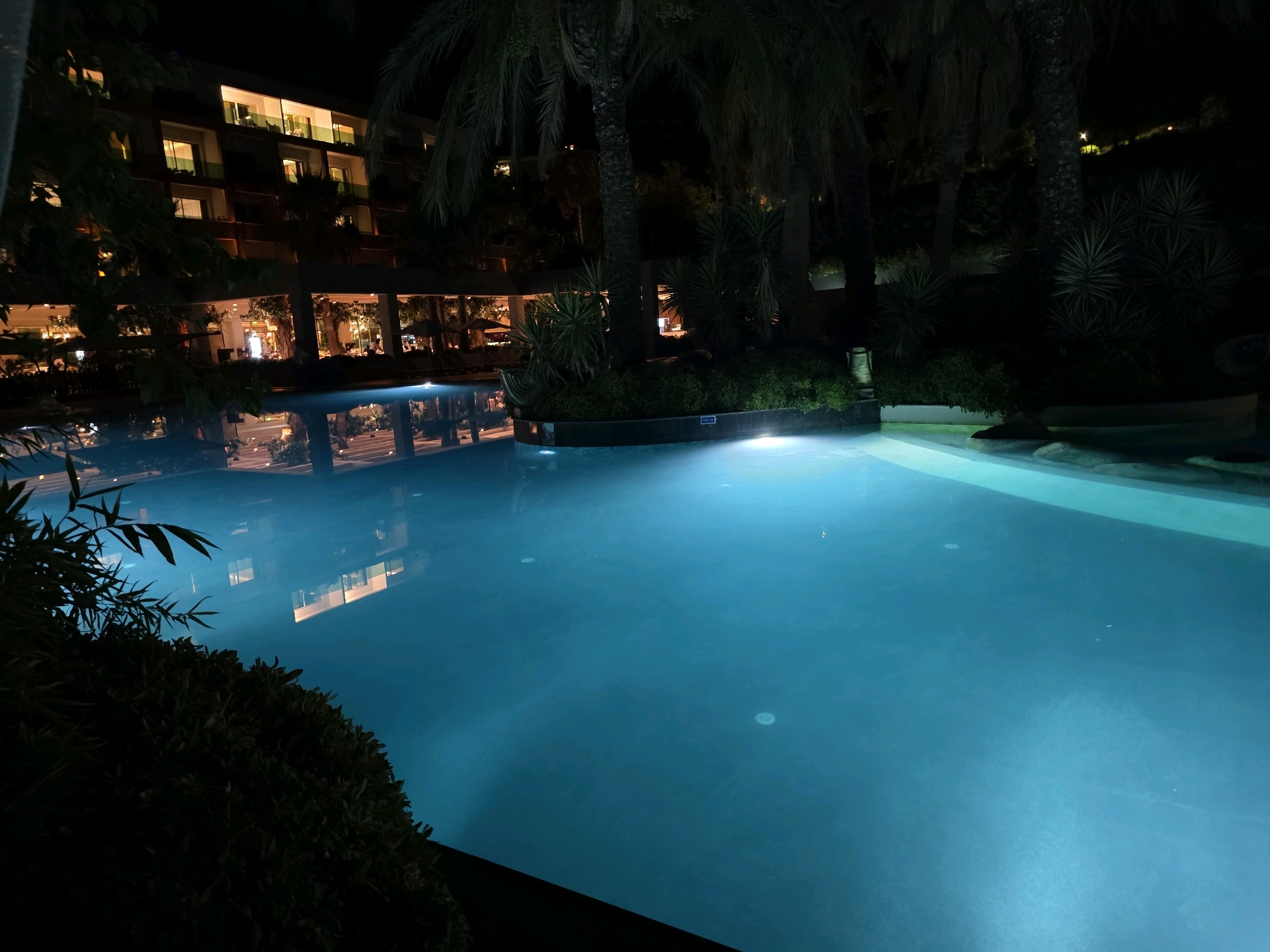 An outdoor swimming pool is lit at night in Bodrum, Turkey. The pool water is a bright, luminous blue, with underwater lights creating a serene glow. Reflections of a multi-story building with illuminated windows are visible on the water's surface. Palm trees and lush greenery surround the pool, creating a tropical atmosphere. The building appears to be a hotel or resort, with warm light spilling from its many windows. A shadowy foreground shows the edge of the pool and some vegetation, adding depth to the scene. The overall ambiance is peaceful and inviting, characteristic of a nighttime resort experience.