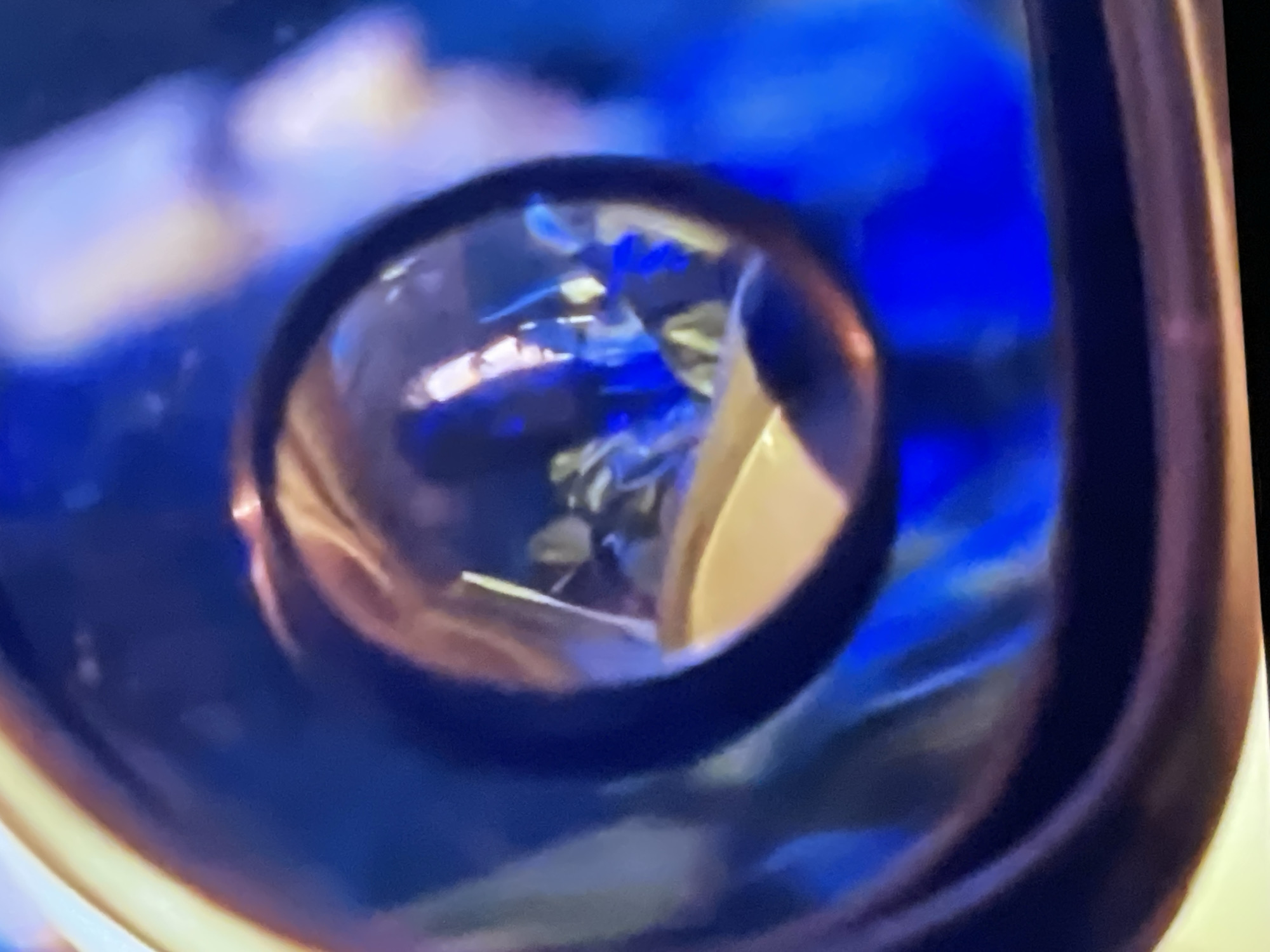 The image shows a reflection in a circular mirror or lens, possibly a rearview mirror or a camera lens. The reflection reveals a person's profile, with a dominant blue and yellow color palette in the background. The person appears to be looking to the left, with their face partially illuminated. The details are blurry due to the nature of the reflection and the image quality. The context of Pattaya City, Thailand, does not appear to be directly discernible from the visual information within the image itself.
