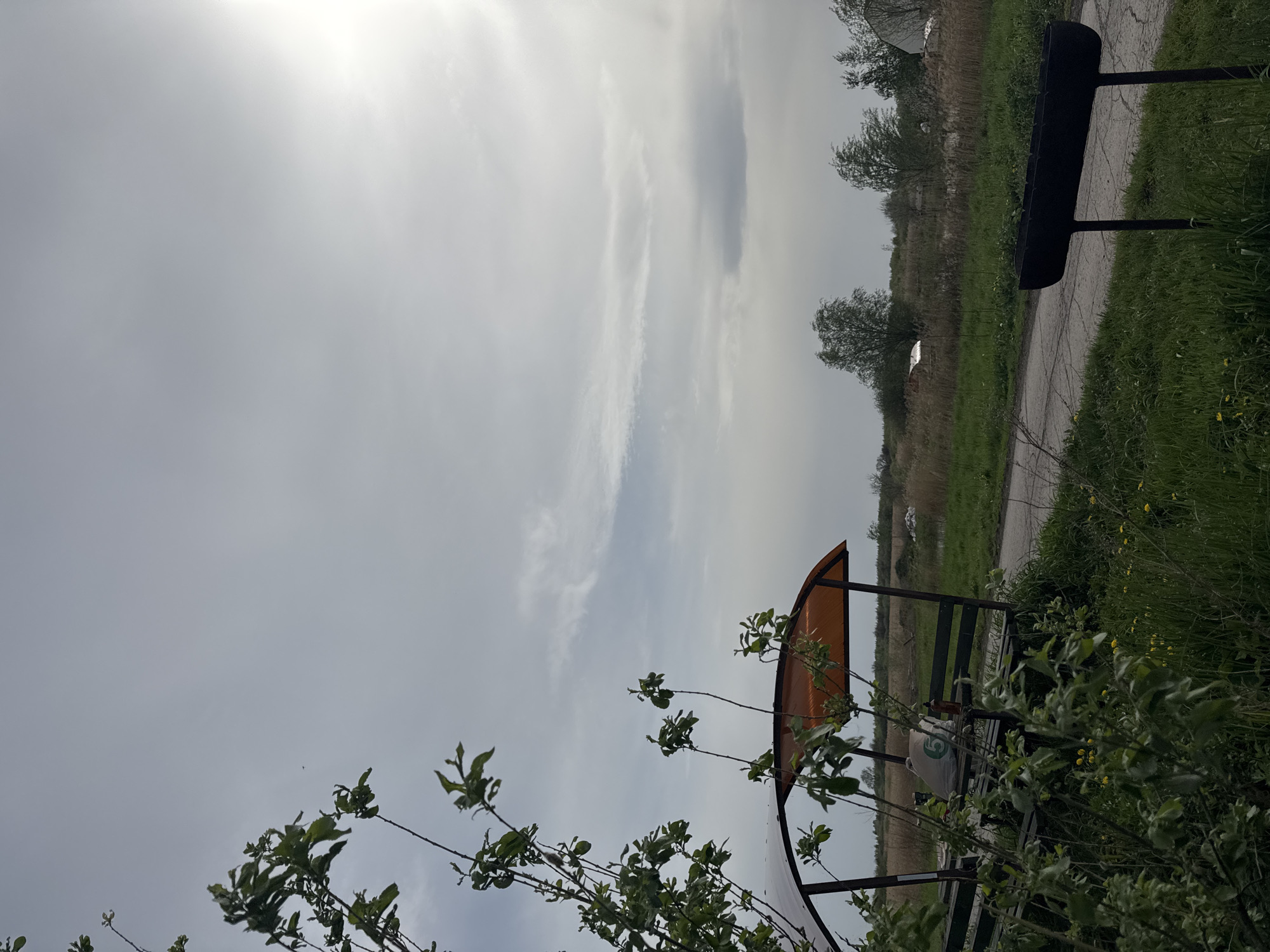 The image shows an outdoor scene, likely a park or rural area, under a cloudy sky. In the foreground, there are branches of a tree or bush with green leaves partially obscuring the view. Further into the scene, there is a covered bench or shelter with an orange roof. Beyond the bench, there is a path or road leading into a field or grassy area. A black structure is visible at the top right of the image, possibly part of a bench or other outdoor fixture. The location is unspecified beyond Unknown, Unknown. There are no people visible in the image.