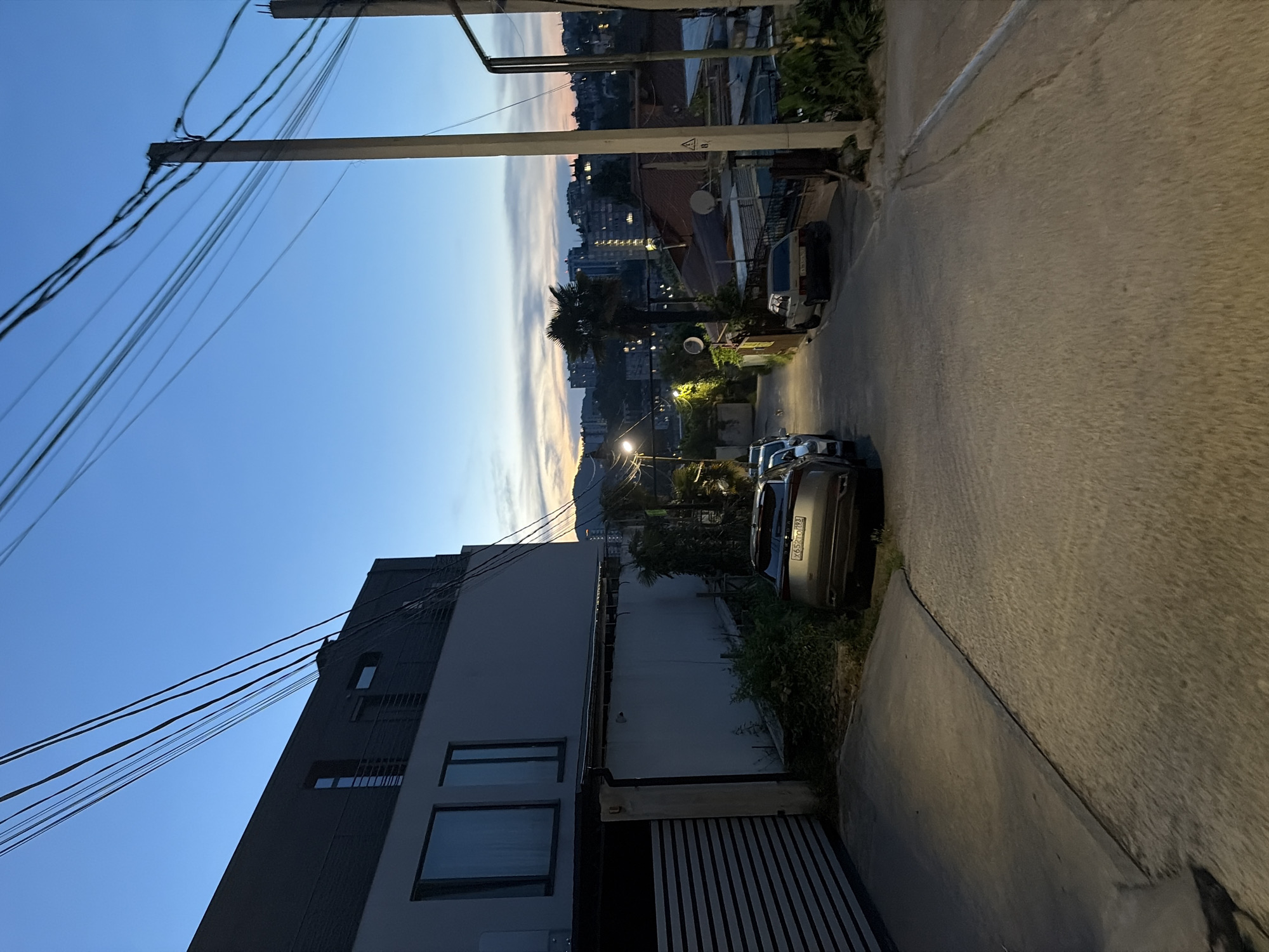 A twilight view of a narrow, paved street in Sochi, Russia, with buildings and a clear sky. The street slopes downwards from the foreground. To the left, a modern, grey building with large dark-framed windows is visible, adjacent to a grey striped garage door. Several cars are parked along the right side of the street, including a silver SUV with a visible license plate reading "X652TO 193". Further down the street, other vehicles and more modest buildings with tiled roofs are seen. Trees, including palm trees, are scattered throughout the scene. Electrical wires are prominently strung across the sky, supported by concrete poles. The sky displays a gradient of blue and pink hues, indicating the sun has recently set. The overall impression is a residential urban setting at dusk.