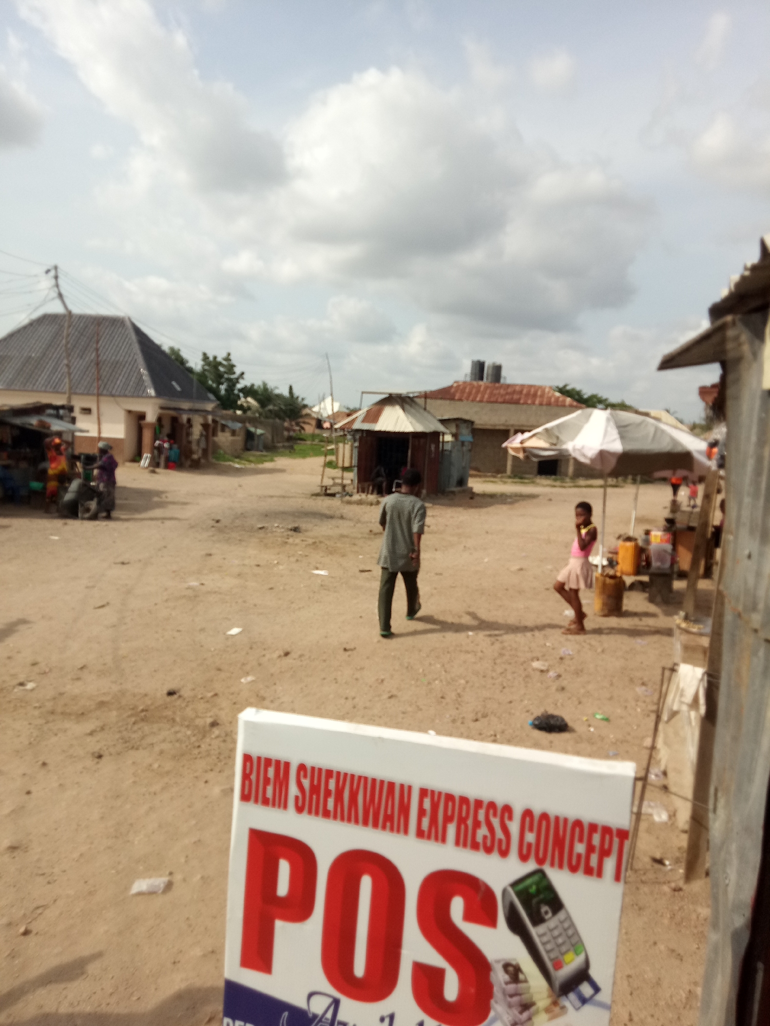 A low-angle, medium shot shows an outdoor, dirt-road scene in a village-like setting. The sky above is overcast with white and gray clouds. In the foreground, a white sign with red lettering reads "BIEM SHEKKWAN EXPRESS CONCEPT POS". Below "POS", the image shows a credit card machine and credit cards. Behind the sign, a man in a grey shirt and green pants walks away from the camera. To the right, a young girl in a pink top and a light-colored skirt stands under a white and gray umbrella next to a stall with containers. In the background, a few buildings with tin roofs line the dirt road, and several people are visible near them. The scene suggests a commercial area or marketplace.