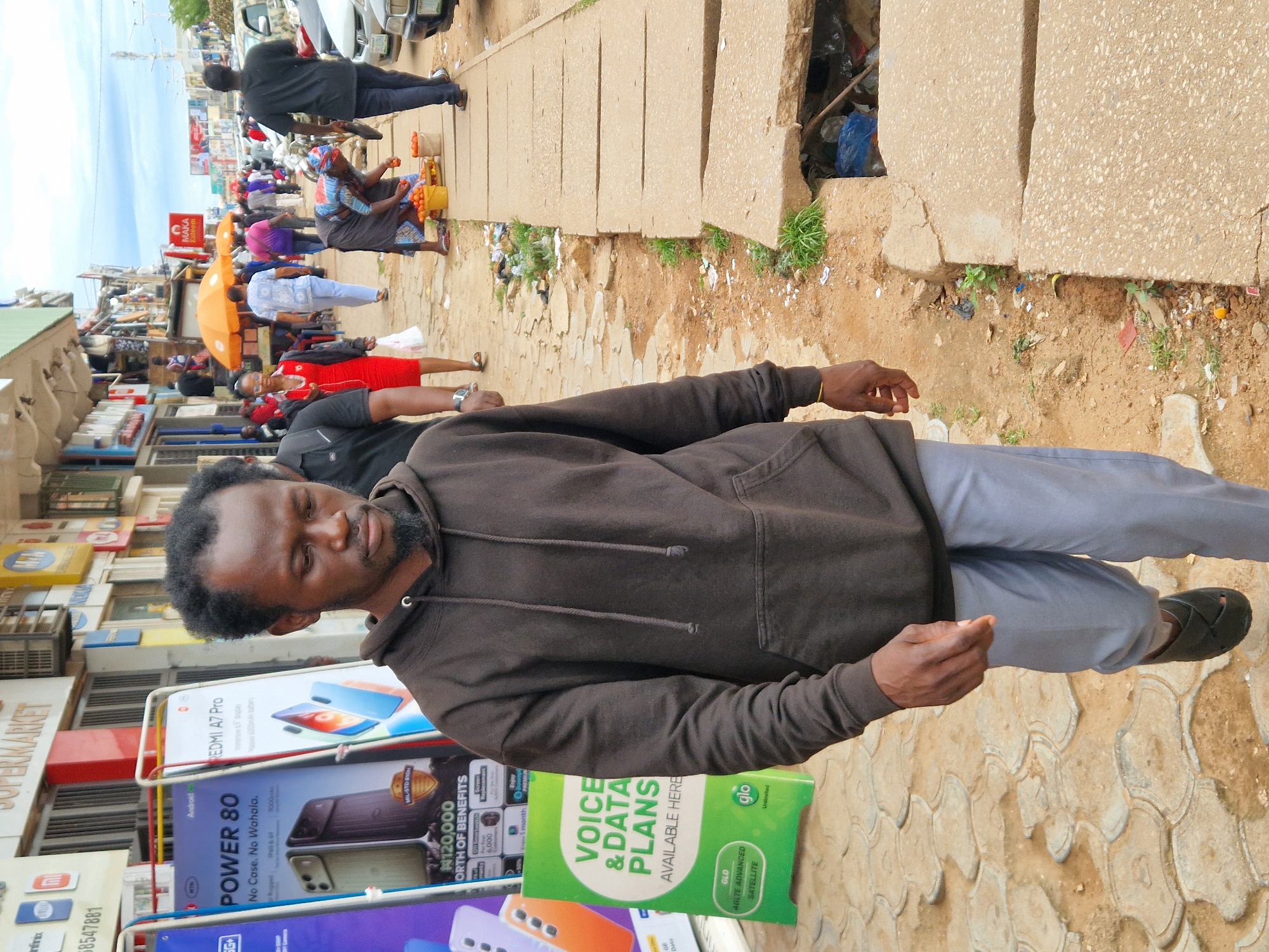 The image depicts a street scene in Jos, Nigeria. In the foreground, a Black man wearing a dark brown hoodie and light grey pants walks along a dirt and paved walkway. He has a short, dark, curly hairstyle and a goatee. To his left, there are large advertisements for mobile phone plans and devices. The street is lined with various shops and businesses, many with colorful signage.
Further down the street, several people are visible. One woman in a bright red dress is walking away from the camera, and another woman is bent over, tending to baskets of oranges. Several orange umbrellas are open, providing shade. Cars are parked along the side of the road. The environment appears to be a bustling commercial area with signs of urban development and some areas with accumulated debris and an open drainage ditch. The sky is overcast.