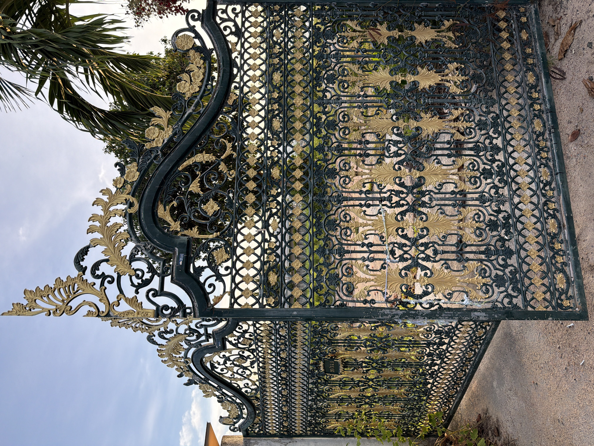 This is a low-angle, vertical shot of an ornate, dark green metal gate with gold accents, seen from the ground level, with the sky and palm fronds visible in the upper left. The gate is elaborately decorated with scrolling acanthus leaves and floral motifs, typical of wrought ironwork. The sunlight catches the gold detailing, highlighting the intricate patterns. Behind the gate, through the openwork, glimpses of foliage and structures can be seen, suggesting a garden or courtyard setting. The overall impression is one of grandeur and artistry. There is no discernible activity or event taking place, and the scene appears tranquil. The time of day appears to be daytime, with a bright sky and dappled sunlight. The location context of Baan Plai Laem, Thailand, suggests a warm climate and possibly a tropical or subtropical environment, which is consistent with the presence of palm trees. There is no visible text in the image.