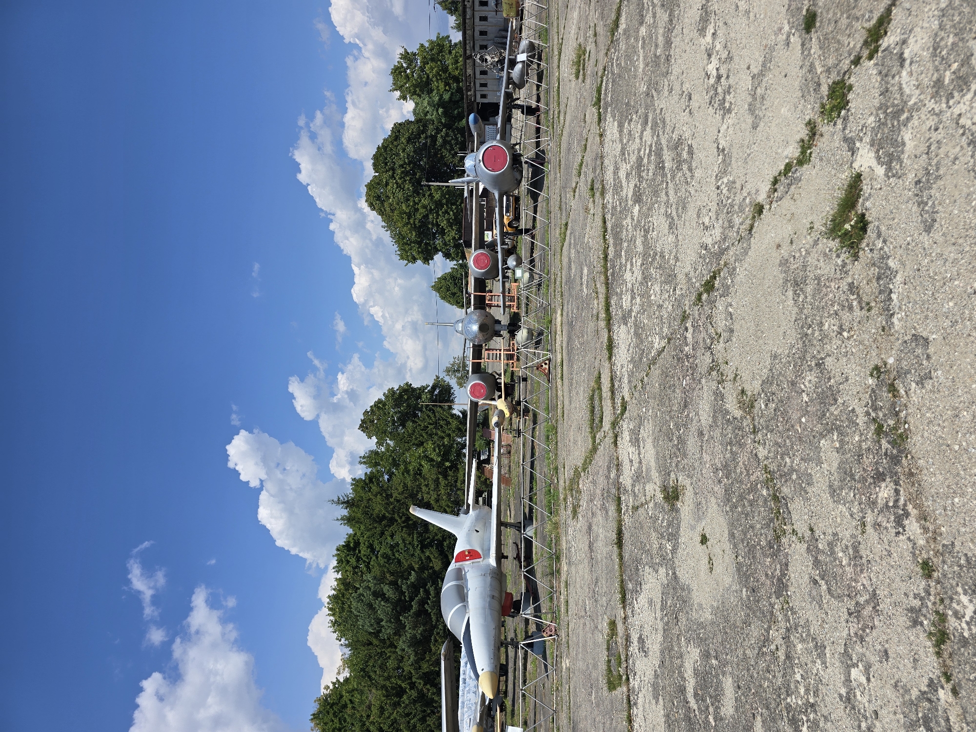 The image shows a sunny day in Pustiměř, Czechia, with a clear blue sky dotted with white clouds. In the foreground, a cracked concrete surface is visible, with patches of dry grass and weeds growing through the cracks. A metal fence separates the concrete area from the rest of the scene. Behind the fence, several vintage aircraft are parked on the tarmac.
The aircraft appear to be in various states of preservation. The closest aircraft to the left is a sleek, white jet with red accents on its tail and engine nacelles. It has a distinctive cockpit canopy and a long, pointed nose. Further down the line, other aircraft are visible, some with visible red circular markings on their engines. The aircraft are lined up in a row, suggesting they are part of an outdoor display or museum.
In the background, a line of lush green trees forms a natural backdrop. Behind the trees, a long, low building with a corrugated roof can be seen, possibly a hangar or storage facility. A yellow vehicle is parked near the building, partially obscured by the trees and aircraft. The overall impression is of a static display of historical aircraft under a bright, sunny sky.