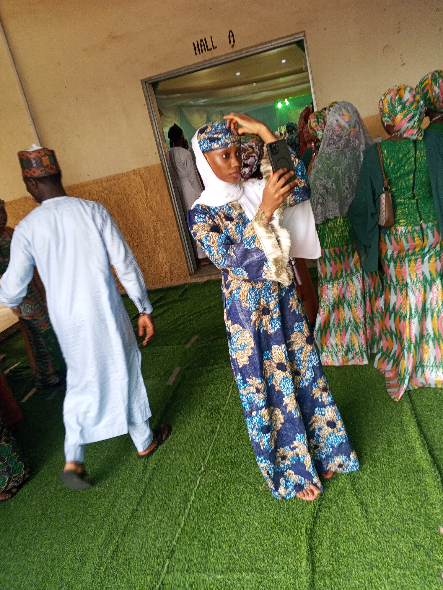 The image depicts an indoor scene at what appears to be a gathering or event, potentially located in Abuja, Nigeria, given the stylistic clothing and context suggested by the signage "HALL A." The foreground features a young woman in a blue and white patterned dress and hijab, holding a smartphone and taking a selfie. To her left, a man in a light blue kaftan and a patterned cap is walking away from the camera, his back partially turned. Behind the woman, several other individuals are visible, mostly women in colorful, patterned attire and headscarves, some of whom are also wearing veils. The floor is covered with artificial green turf, and the walls are a neutral, textured color. The lighting suggests an indoor environment with overhead illumination, creating a bright but somewhat diffused atmosphere. The overall impression is one of a social occasion, possibly a celebration or religious event, taking place within a public hall.