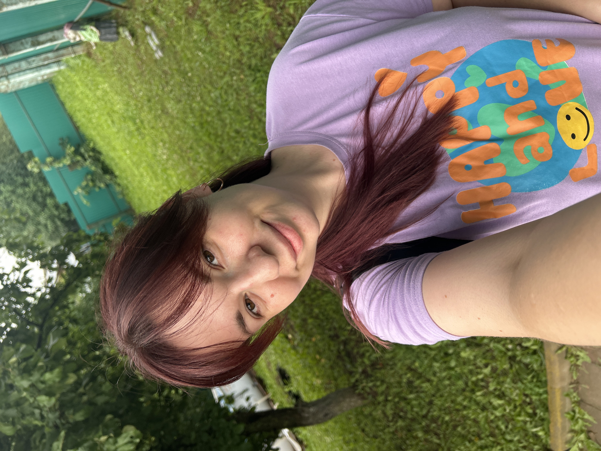 A young woman with auburn hair is taking a selfie outdoors. She is wearing a light purple t-shirt with an orange and blue graphic that says "HAPPY and LOVE" with a smiley face. She is smiling and looking at the camera. The background shows a green lawn, a tree, and a green fence. There is a small statue of a person in the background. The scene appears to be in a residential backyard. The location is stated to be in the city of Fokino, Russia.