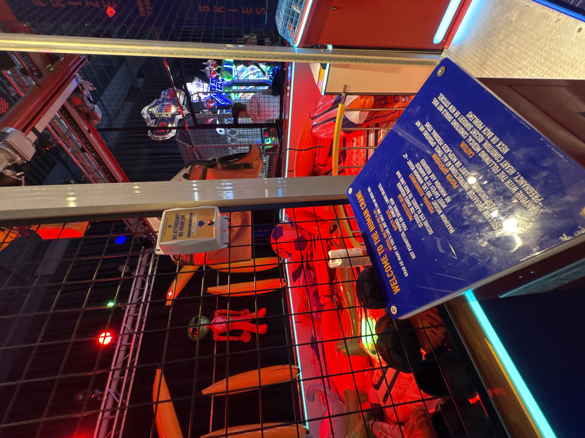 The image depicts the interior of an arcade or amusement venue, likely in Panama City Beach, United States of America, based on the context of similar venues in that location. The focal point is a large, blue sign for "THE HUMAN CRANE" game, which is partially visible and displays safety rules and operational instructions. The game itself appears to be a claw or crane machine designed to pick up prizes.
In the background, through a black metal grate, various arcade games and attractions are visible, illuminated by vibrant red and blue lights. Some of these include digital scoreboards showing numbers like "50" and "25.0". Several people are present within the venue, though their faces are not clearly distinguishable due to the lighting and distance. One individual in a white shirt is operating one of the games. In the foreground, a stuffed alien figure in red clothing is suspended, presumably as a prize or part of the game's display. The overall environment is characterized by bright, artificial lighting and the typical clutter of an entertainment arcade.