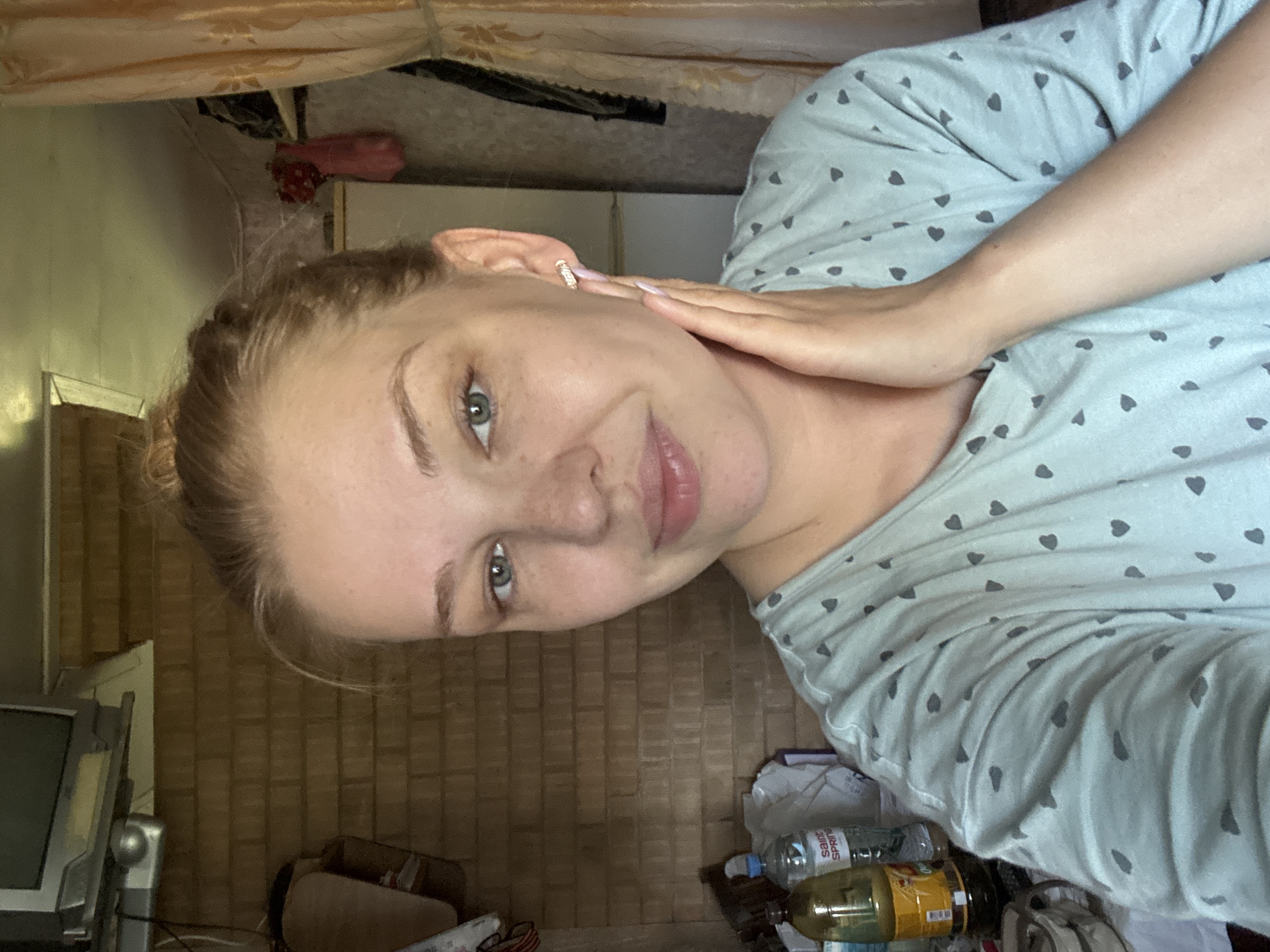The image shows a young woman with blonde hair in a bun, wearing a light blue t-shirt with a heart pattern. She is looking at the camera with a slight smile, her right hand touching her chin. She is wearing small, sparkly earrings. The background appears to be indoors, with a brick wall, a television set mounted on the wall, and some items on shelves. There are also bottles of liquid and bags visible in the background, suggesting a cluttered or storage area. The lighting suggests it may be daytime. The location of Дунилово, Russia, is mentioned in the prompt, but there are no specific visual cues in the image to confirm this location.