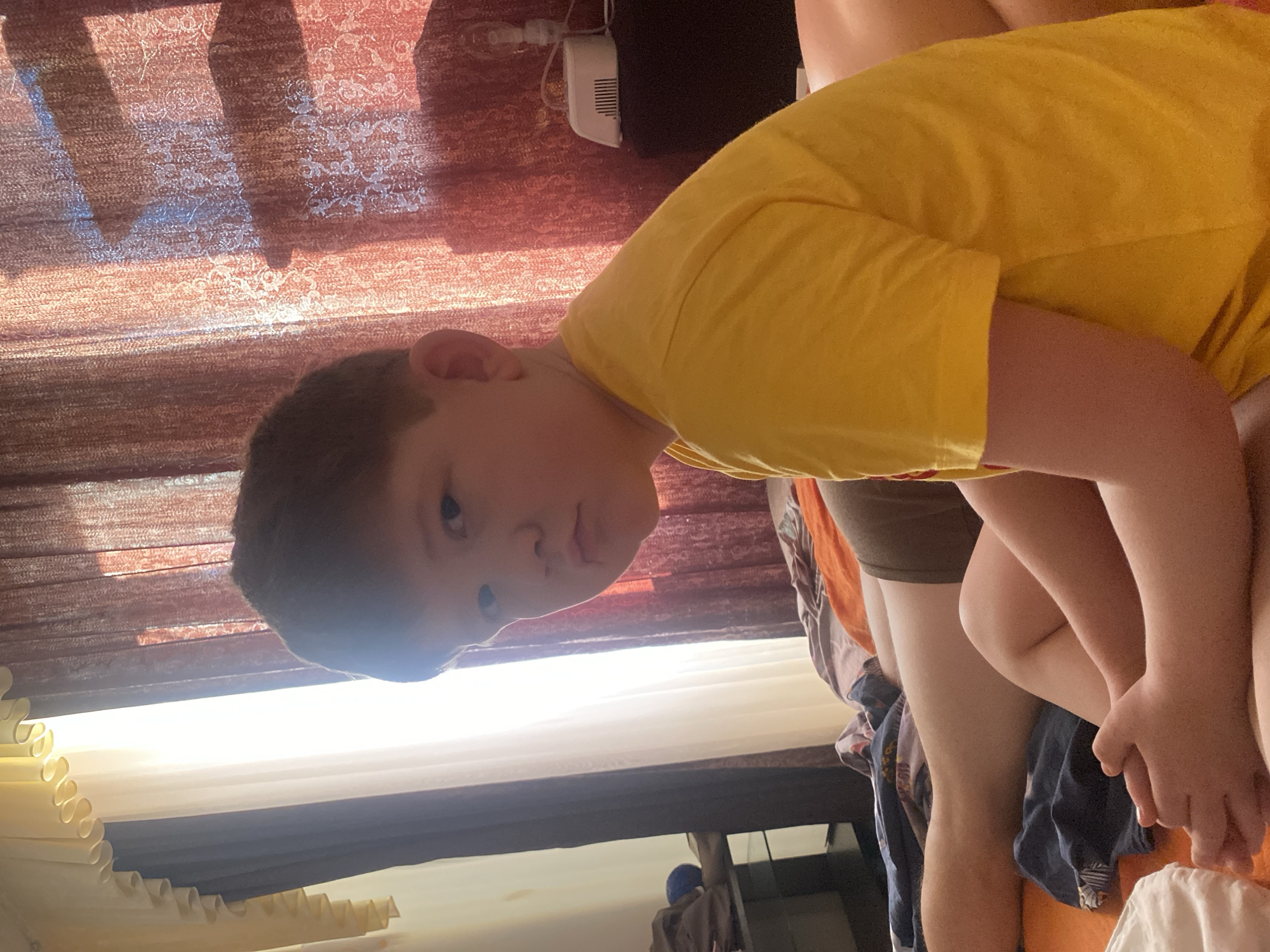 A medium shot photograph shows a child wearing a yellow shirt, looking directly at the camera. The location is a room in what is potentially a residence in Chaikovsky, Russia. Behind the child is a window with partially drawn curtains, a section of decorative wallpaper, and a piece of electronic equipment.