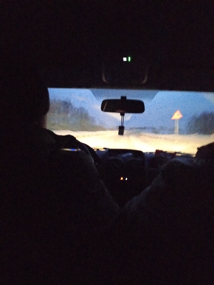 The image displays an interior perspective from a vehicle, facing forward through the windshield, under low-light conditions within Krasnoyarsk, Russia. The silhouette of an individual is partially visible in the driver's seat, viewed from behind, with their right arm resting on the steering wheel, indicating vehicle operation. The dashboard exhibits two illuminated amber indicator lights. An overhead console above the windshield features two distinct green illuminated indicators. A rearview mirror is centrally positioned, with a dark, rectangular object suspended from it. The windshield surface shows numerous small, illuminated specks, suggesting precipitation or condensation. Outside the vehicle, headlights illuminate a dirt or snow-covered road extending into the distance. On the right side of the road, a yellow triangular warning sign with a red border is present, though its specific symbol is not discernible. Dark vegetation or trees line the road's periphery against a dimly lit sky.