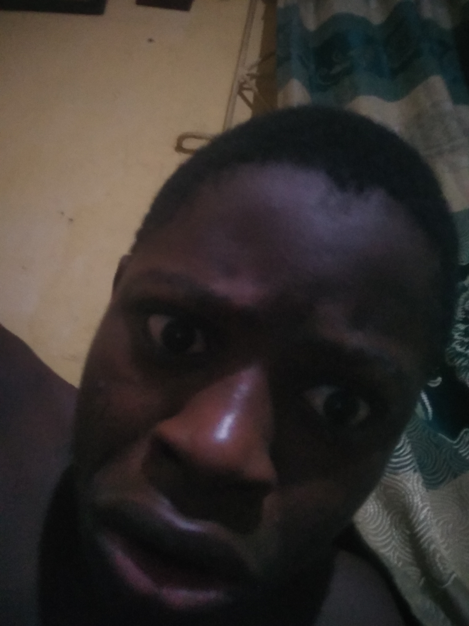 A close-up, low-angle photograph shows a dark-skinned man's face. His expression appears tense, with wide eyes. The background is blurry and dark. A patterned curtain is visible on the right side of the image. The location is presumed to be Shangisha, Nigeria.