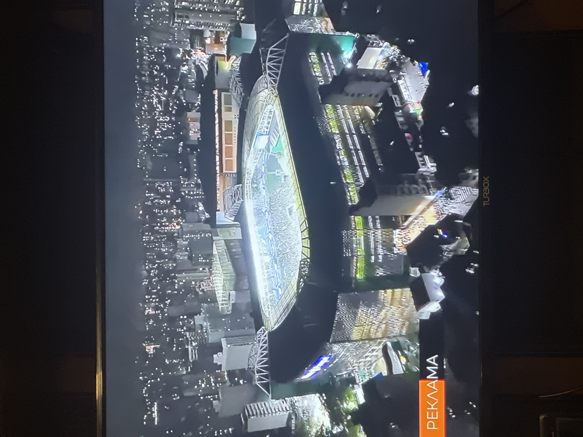 This is an aerial night shot of a stadium in a city. The stadium is lit and appears to be filled with spectators. The surrounding area is densely populated with buildings and streetlights. The word "РЕКЛАМА" (which translates to "advertisement" in Bulgarian) is visible on an orange banner in the lower left corner of the screen, suggesting this is a broadcast. A "TURBOX" logo is present at the bottom center of the screen, likely indicating the television provider. The city's skyline is visible in the background, characterized by numerous illuminated windows in the buildings. The specific location of Berkovitsa, Bulgaria, is not discernible from the image.